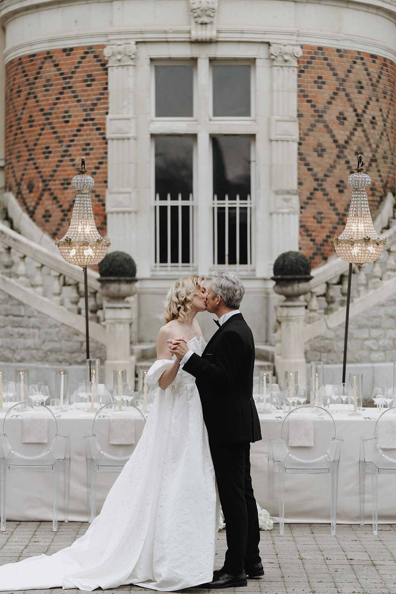 Bride in off-shoulder ballgown and groom in black tuxedo kiss on chateau terrace with reception table behind them