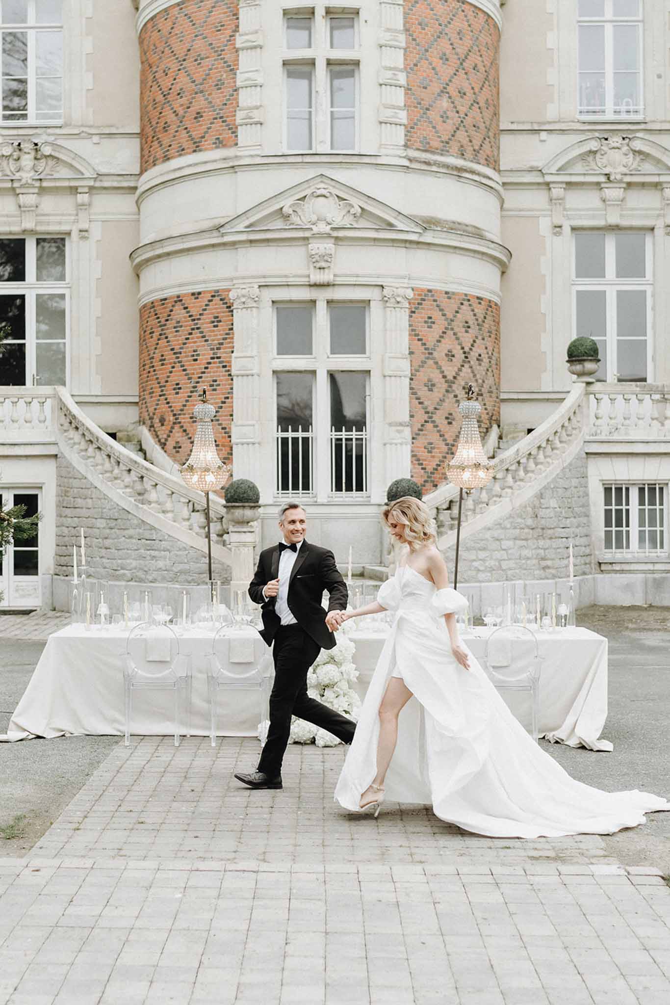 Playful couple posing before ghost chair table with crystal chandeliers and ornate chateau staircase