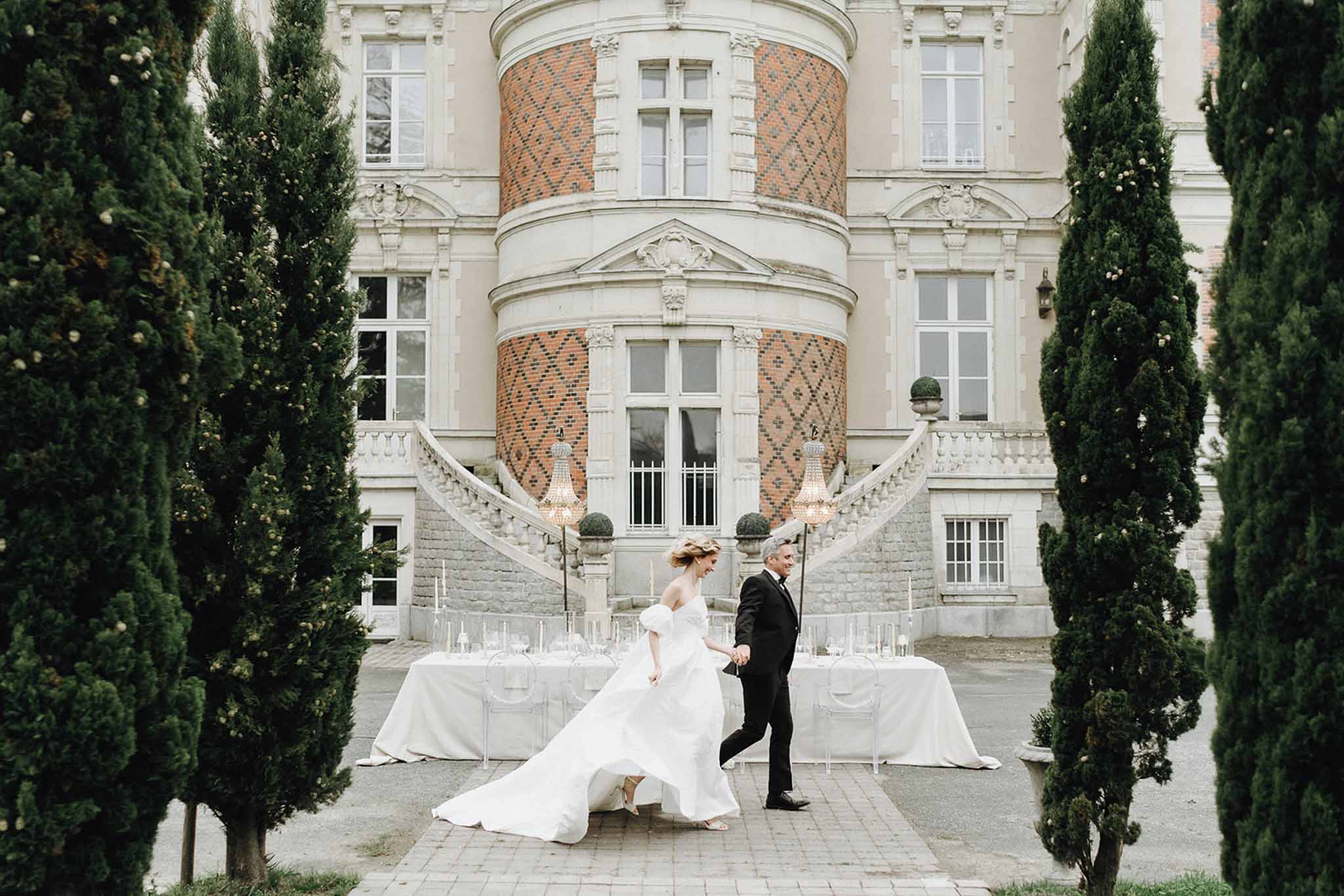 Couple walking across cobblestone courtyard past ghost chair reception table at base of chateau double staircase