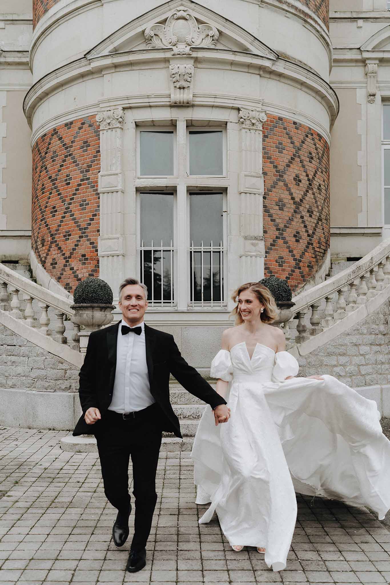 Bride and groom walking hand-in-hand on cobblestone terrace in front of chateau with brick tower and topiary