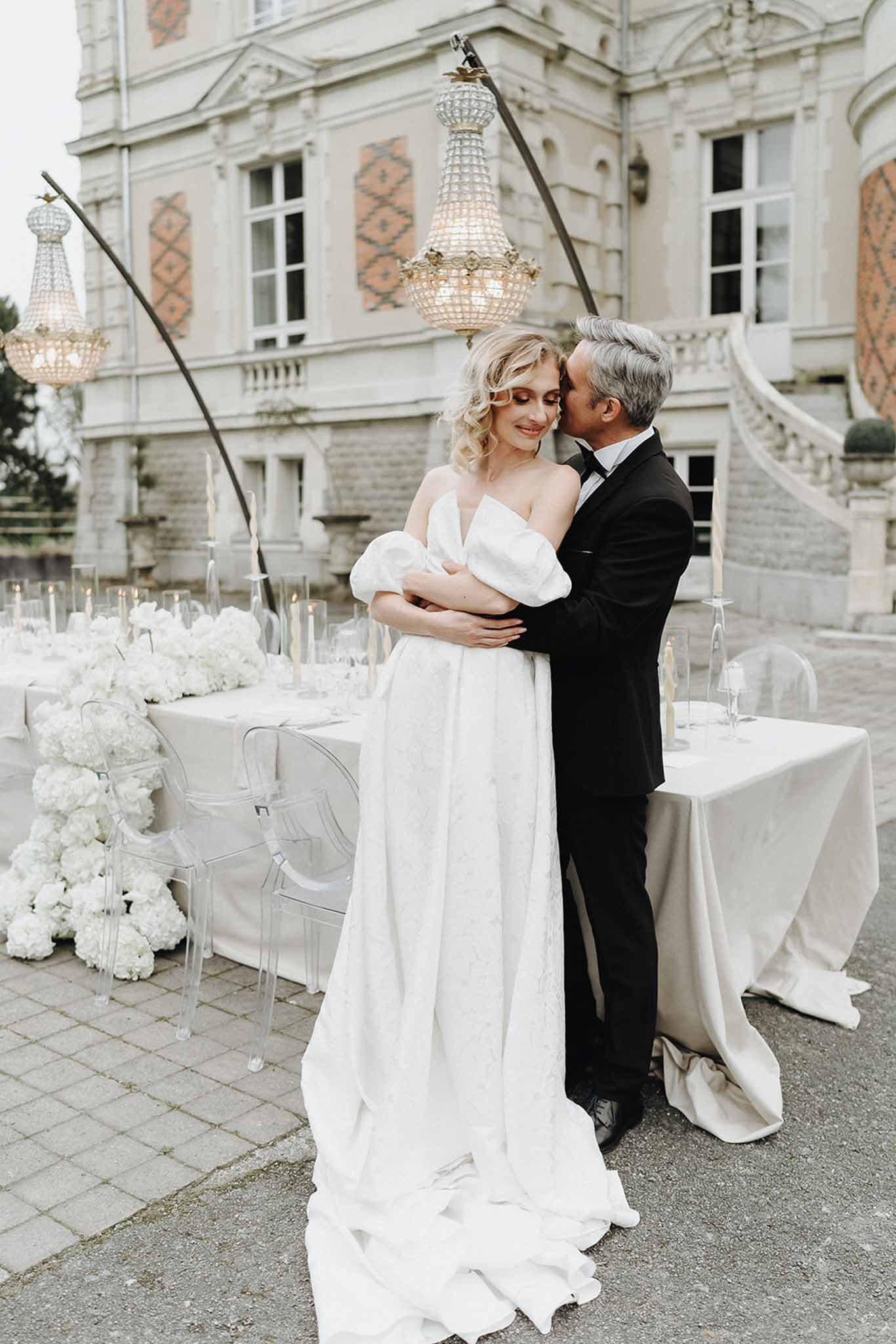 Couple embracing before hydrangea table with suspended chandeliers and brick chateau with stone staircase