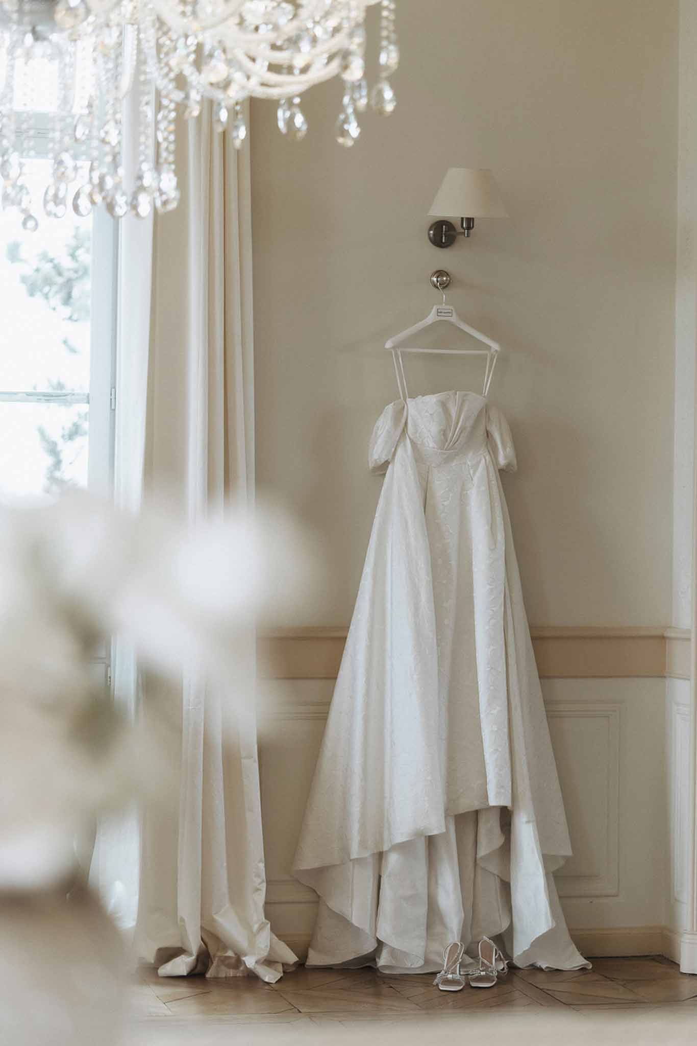 Ivory brocade off-shoulder wedding gown hanging from wall sconce with silver heeled sandals on parquet floor below