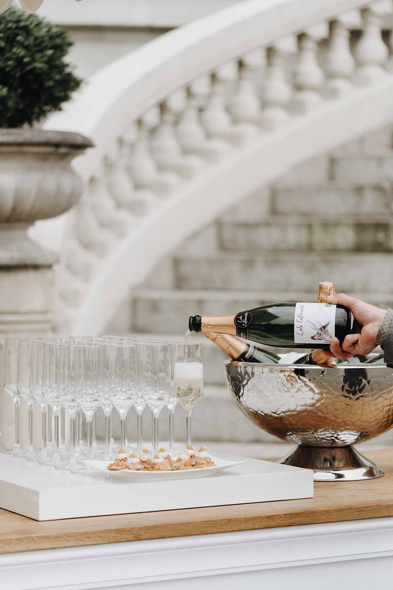 Champagne being poured from silver ice bucket into crystal flutes with canapes on marble tray at chateau terrace