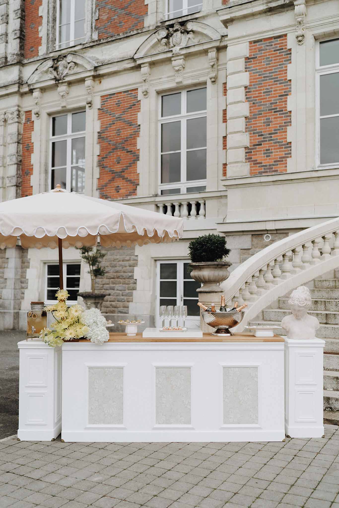 White paneled cocktail bar with scallop umbrella, hydrangeas, and copper bucket at chateau courtyard