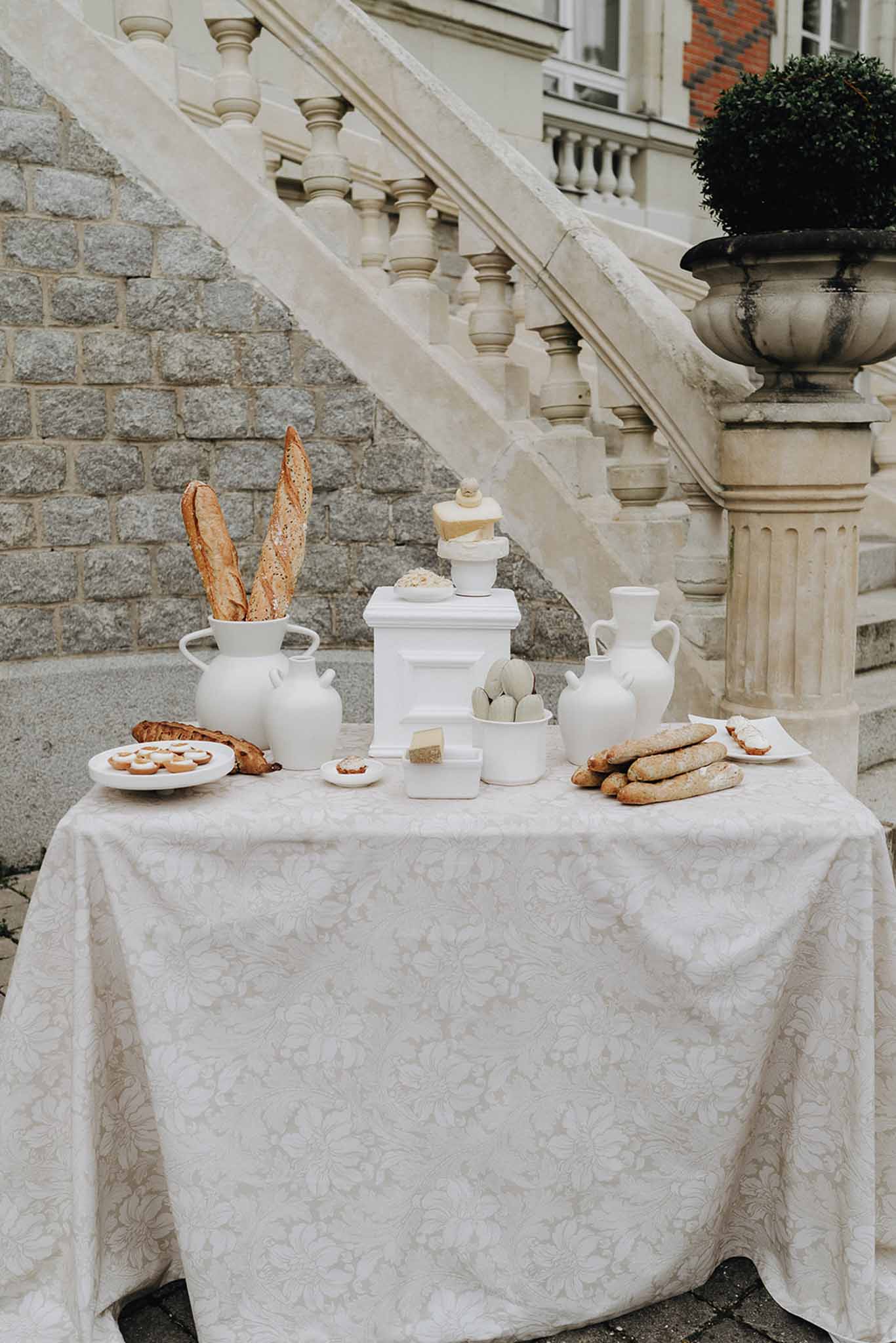 Welcome food display with white ceramic vessels baguettes macarons and pastries on jacquard linen at chateau staircase