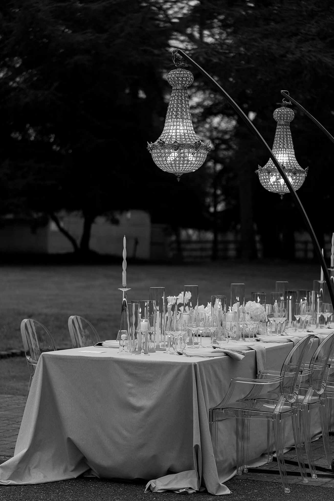 Black and white long table with ghost chairs, hurricane candles, and crystal chandeliers on standing arches