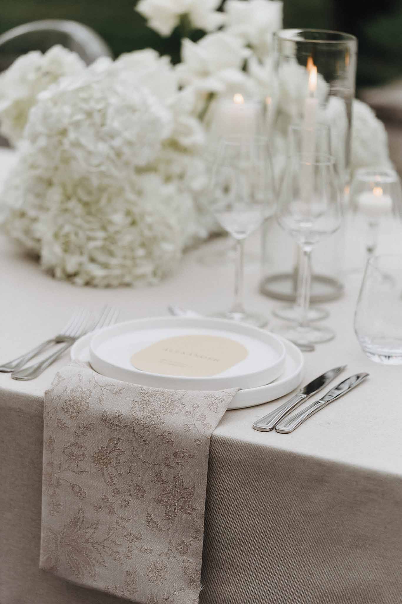 Wedding place setting with stacked white plates, cream place card, crystal glasses, and white hydrangea centerpiece