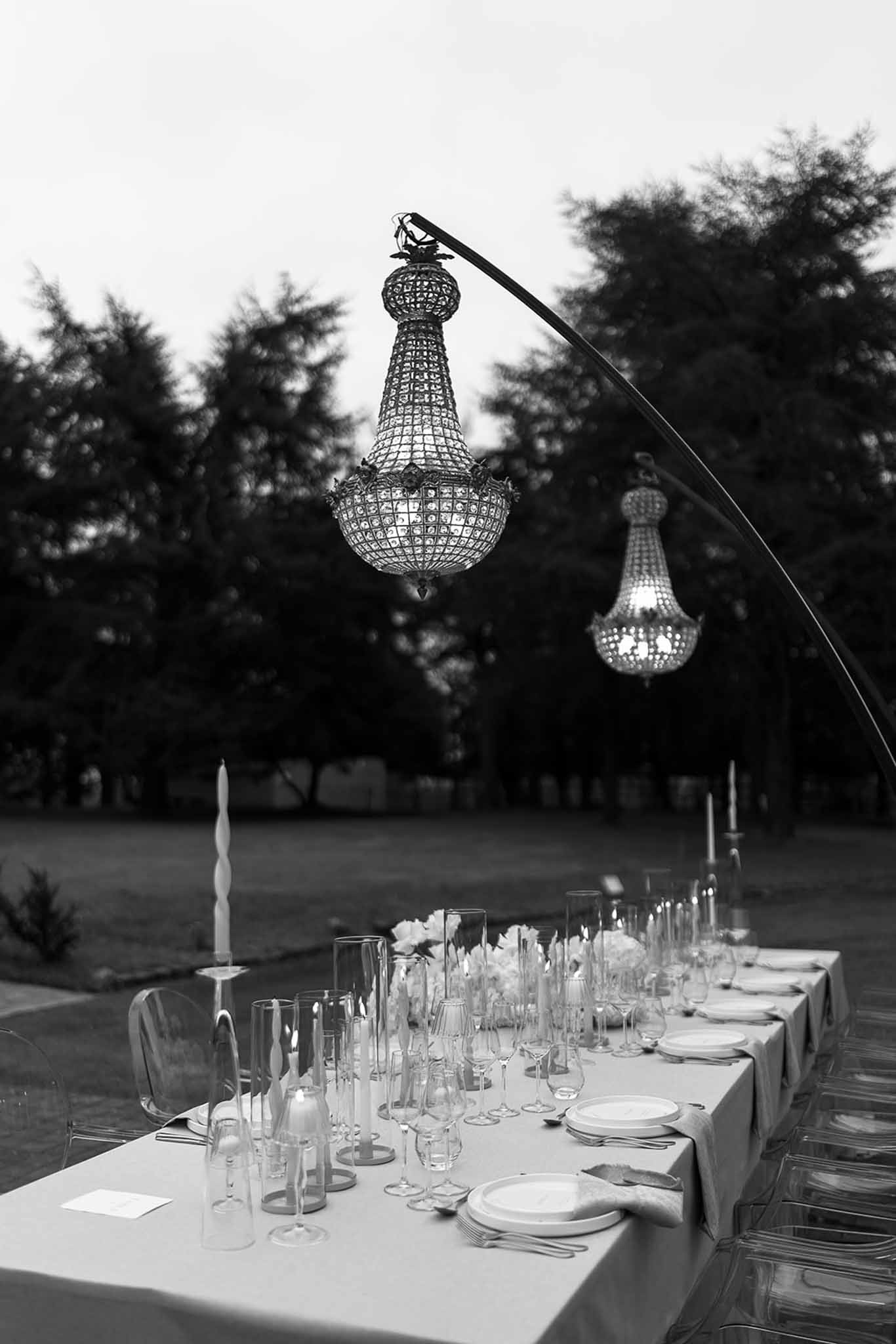 Black and white photo of outdoor reception table with crystal chandeliers ghost chairs and taper candles at dusk