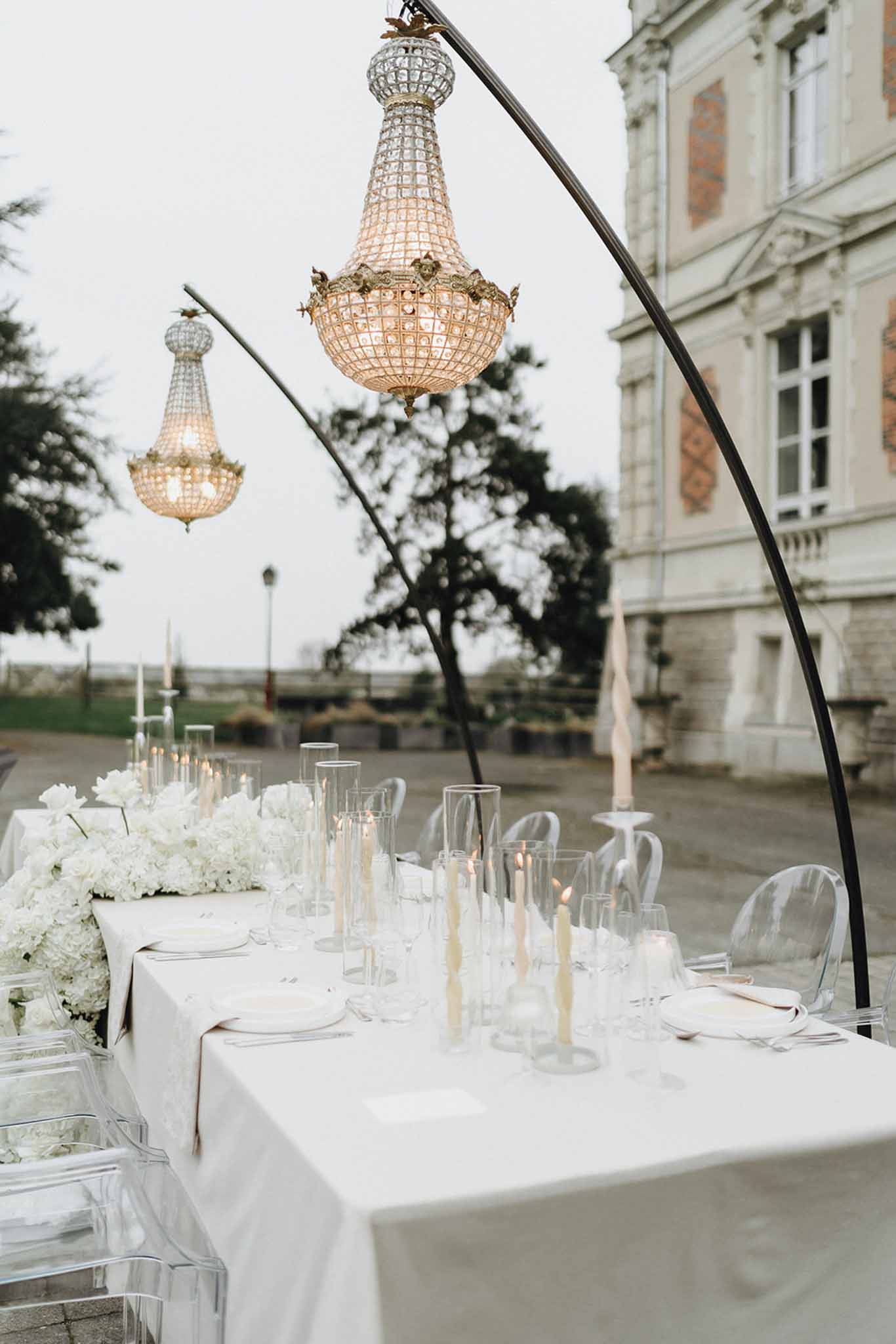 Long reception table with white hydrangea runner, lit taper candles, and hanging crystal chandeliers at dusk