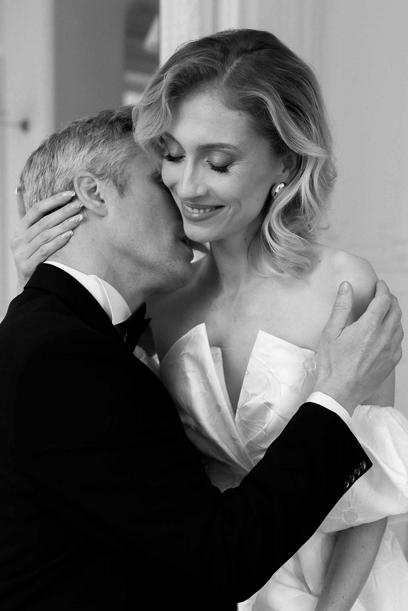 Black and white portrait of groom in tuxedo kissing bride on cheek as she smiles with eyes closed
