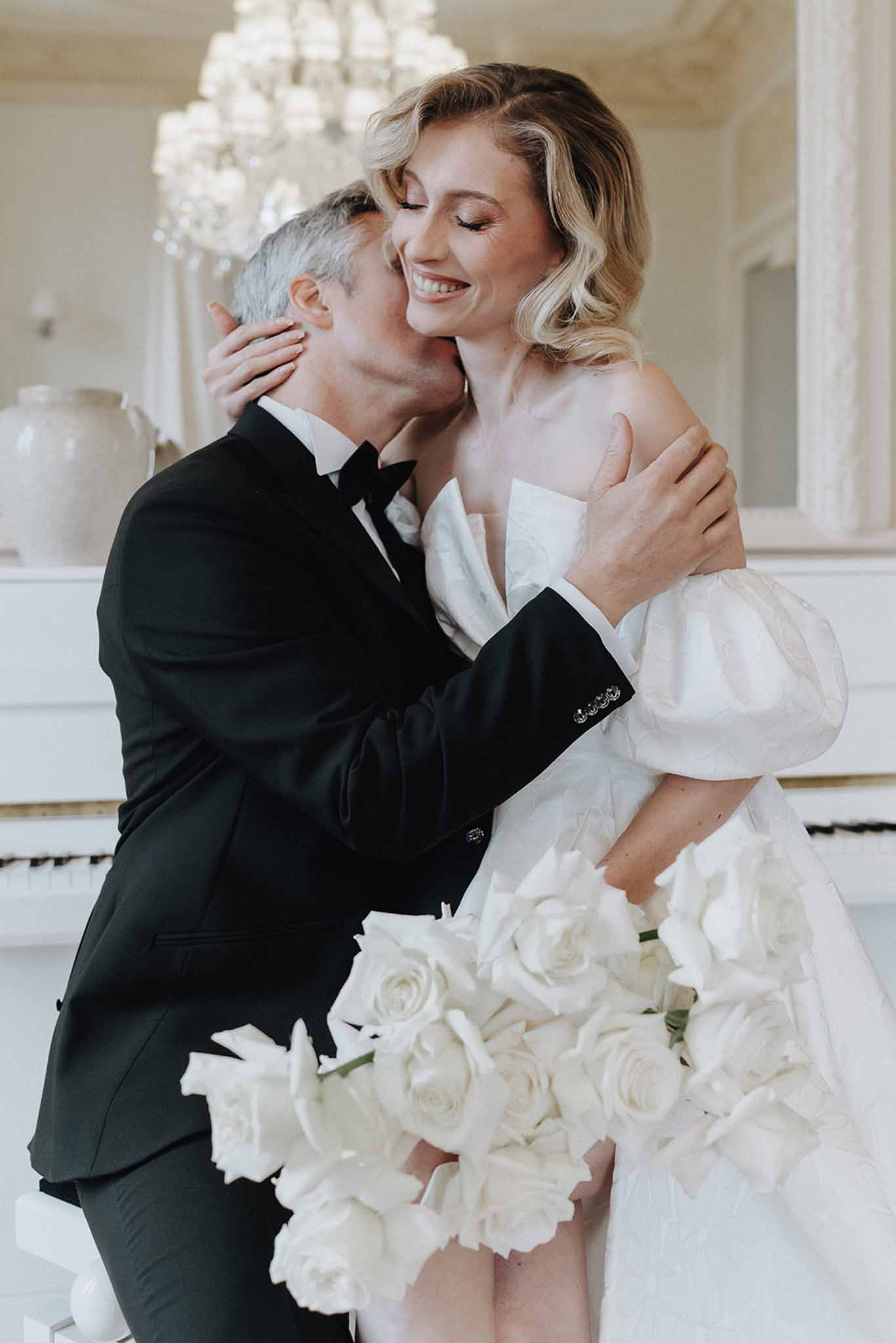 Groom kissing bride on cheek in chateau interior with chandelier and white grand piano