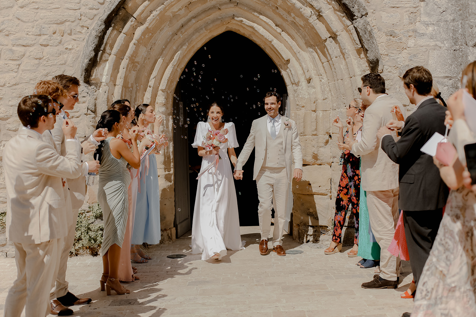 Newlywed couple walking through stone archway with guests blowing bubbles at Mediterranean wedding venue