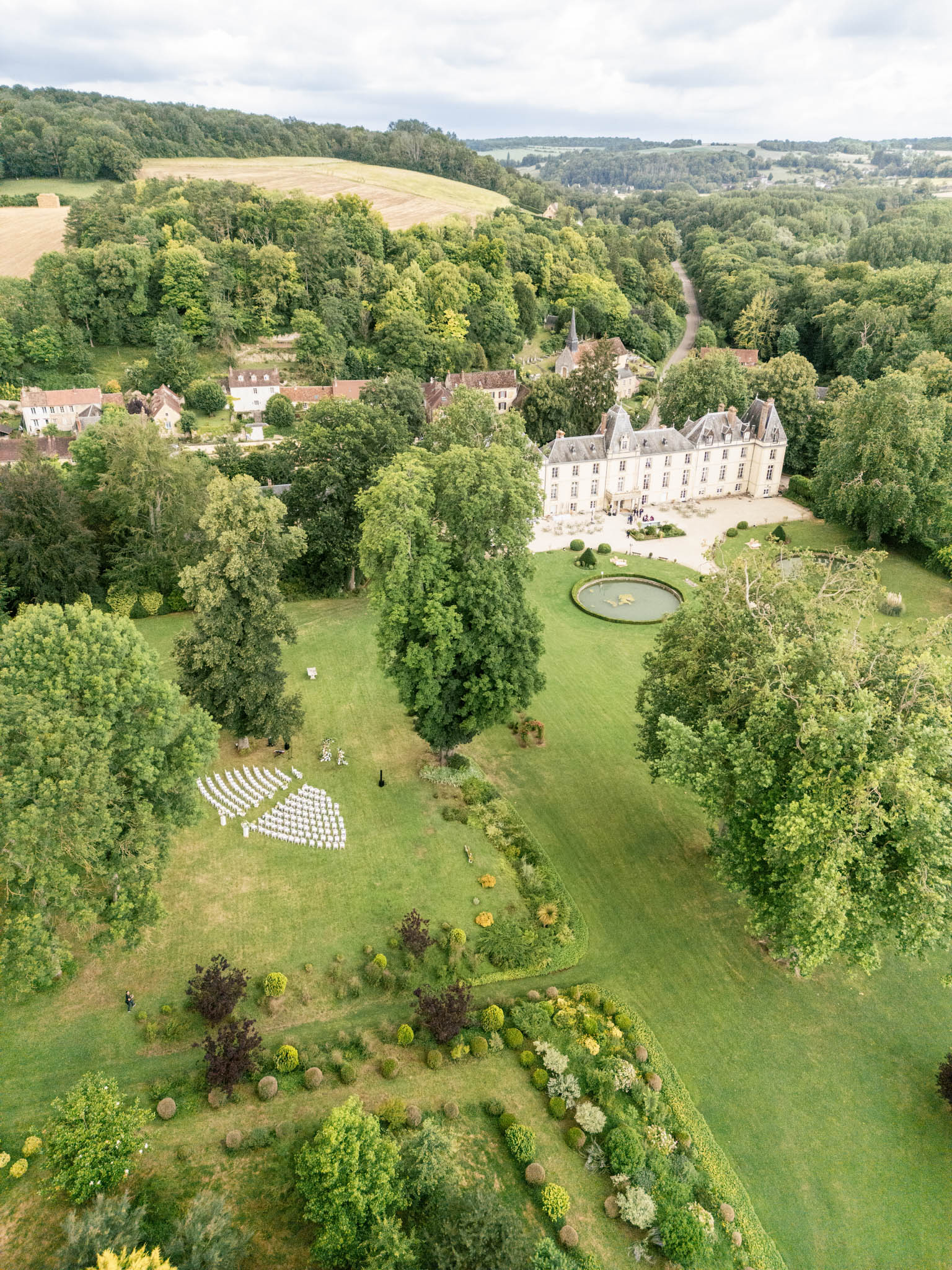 Aerial view of French chateau with outdoor ceremony setup on lawn, ornamental pond, and formal gardens
