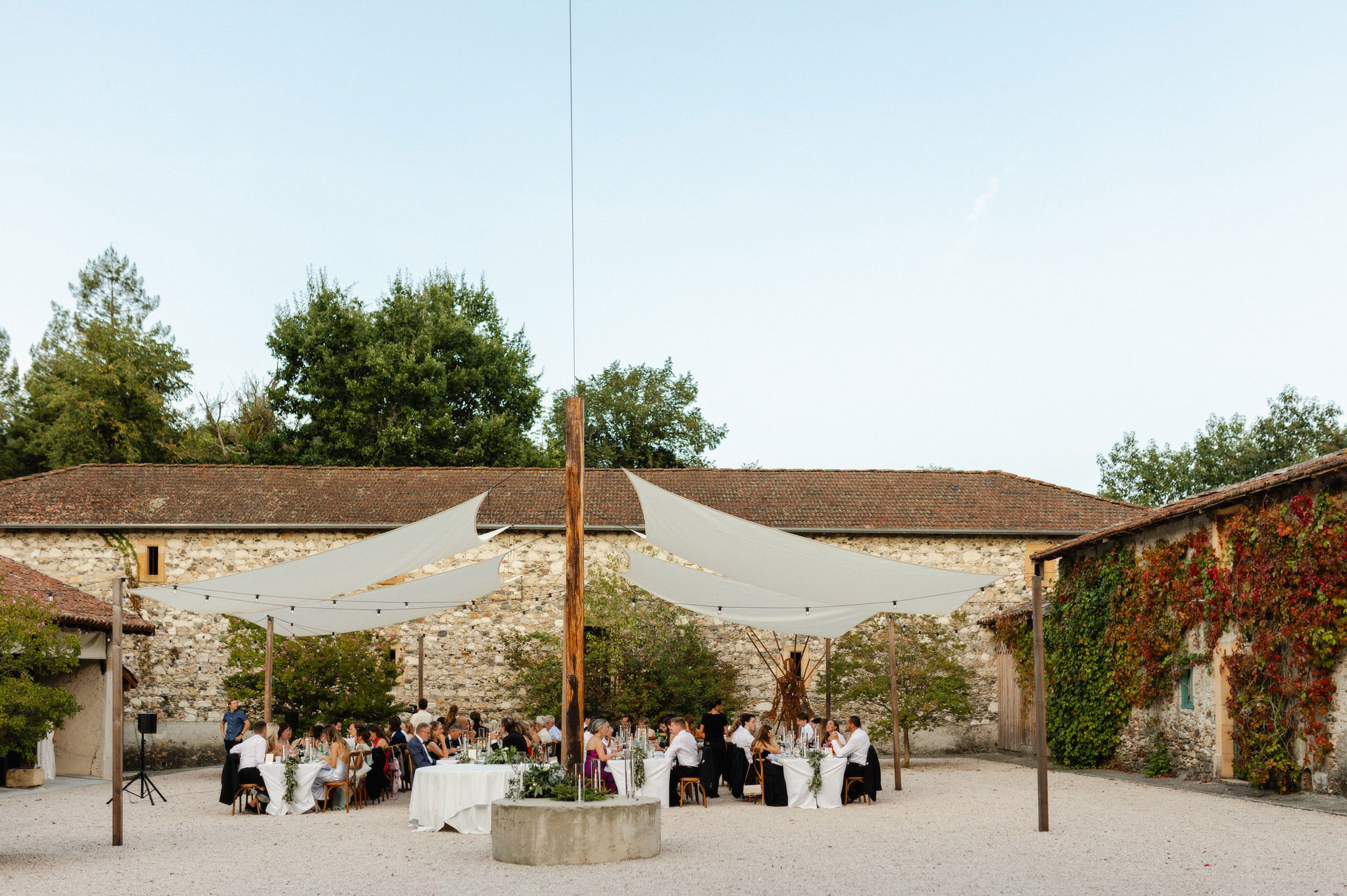 40 guests at round tables under sail shades and festoon lights in stone farmhouse courtyard