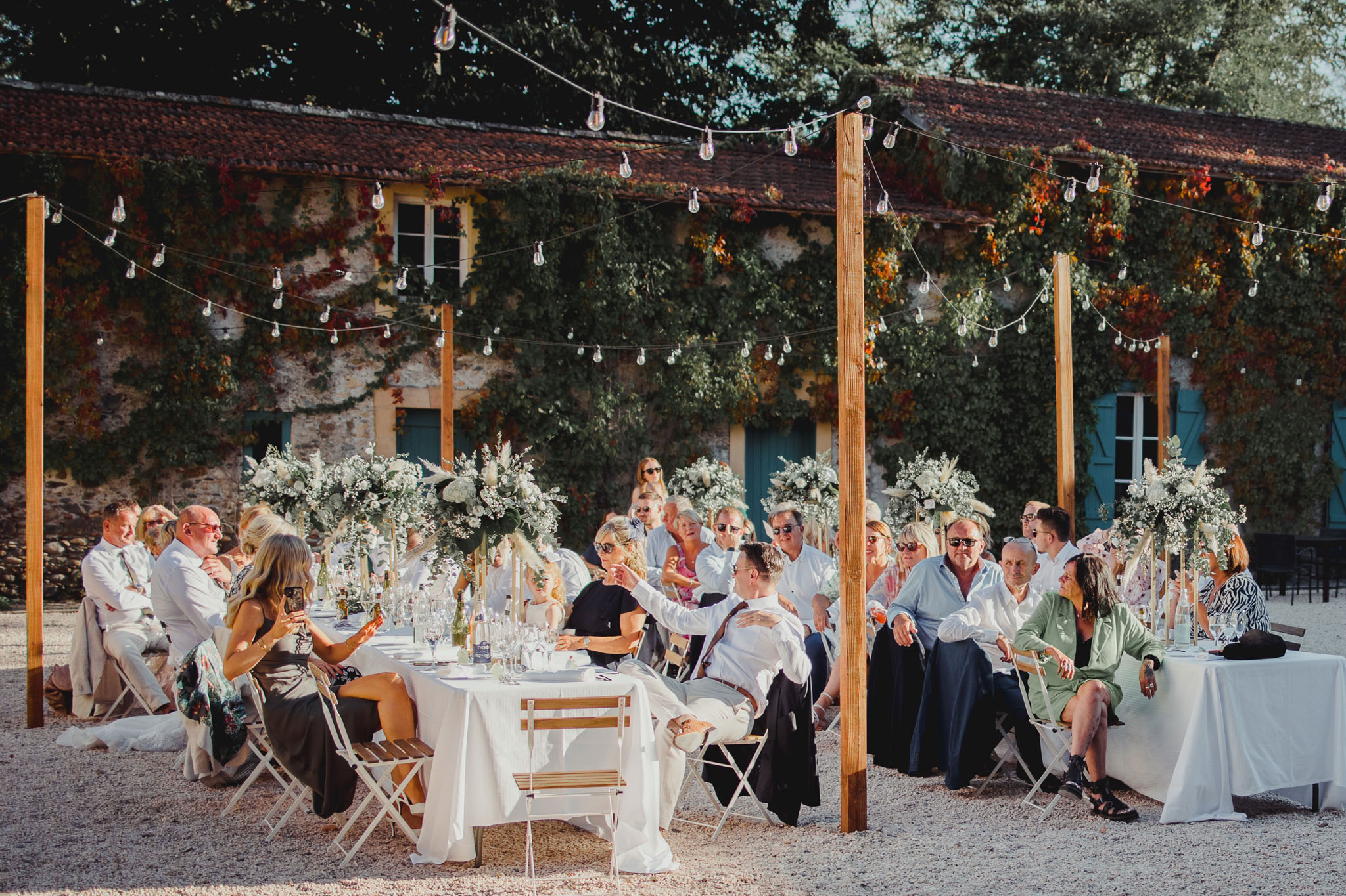 Outdoor reception dinner on gravel courtyard with long tables, festoon lights, and white floral centerpieces