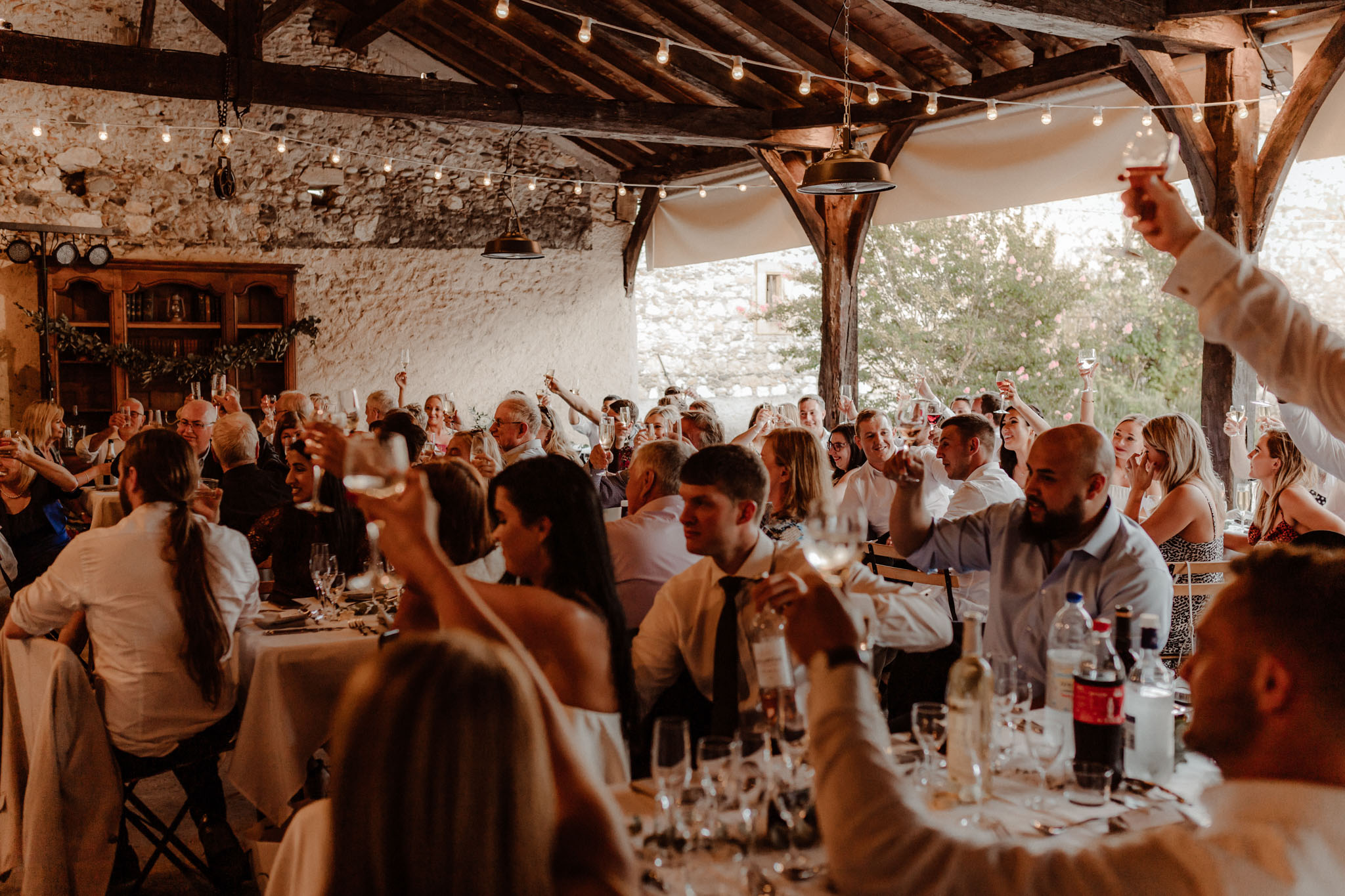 Guests raise glasses in toast inside rustic stone barn with Edison bulb string lights and wooden beams