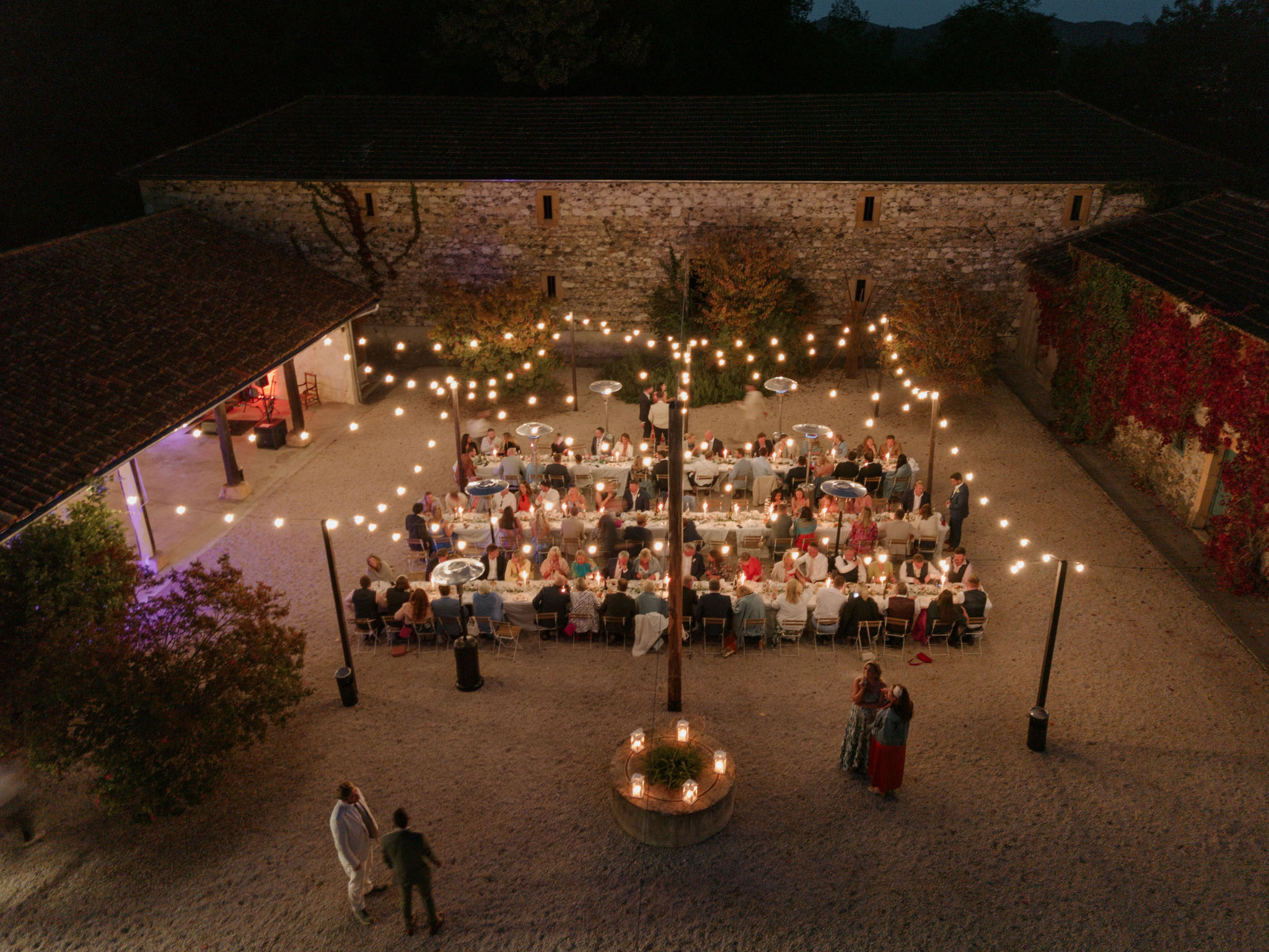 Aerial view of outdoor evening wedding reception in a gravel courtyard with string lights and guests seated at long tables