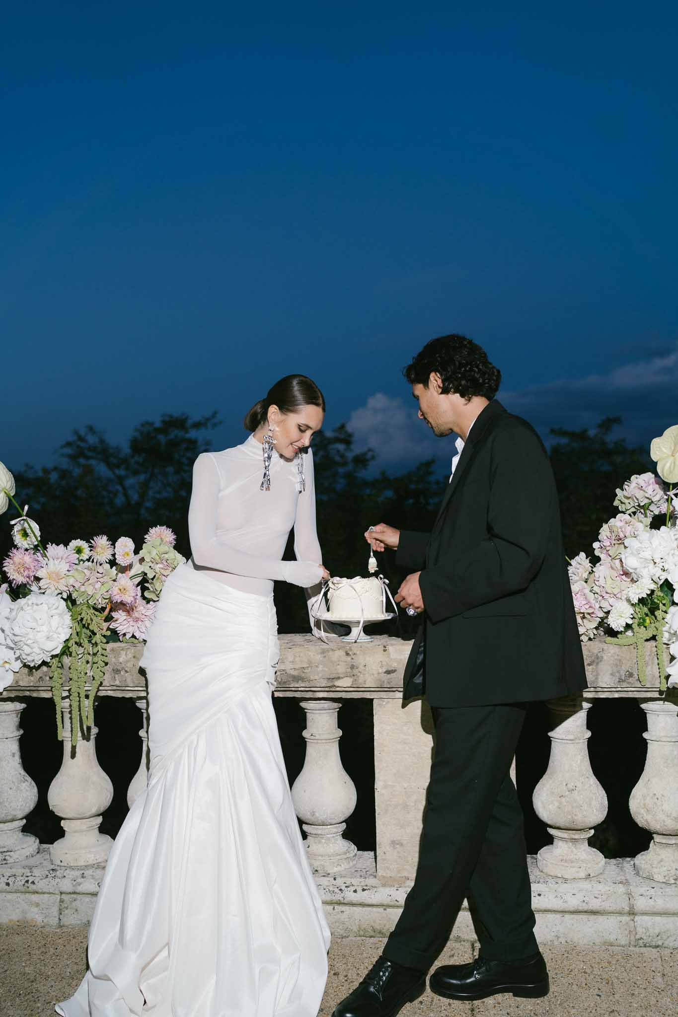 Bride and groom cutting single-tier white cake on chateau terrace at dusk with blush dahlia arrangements