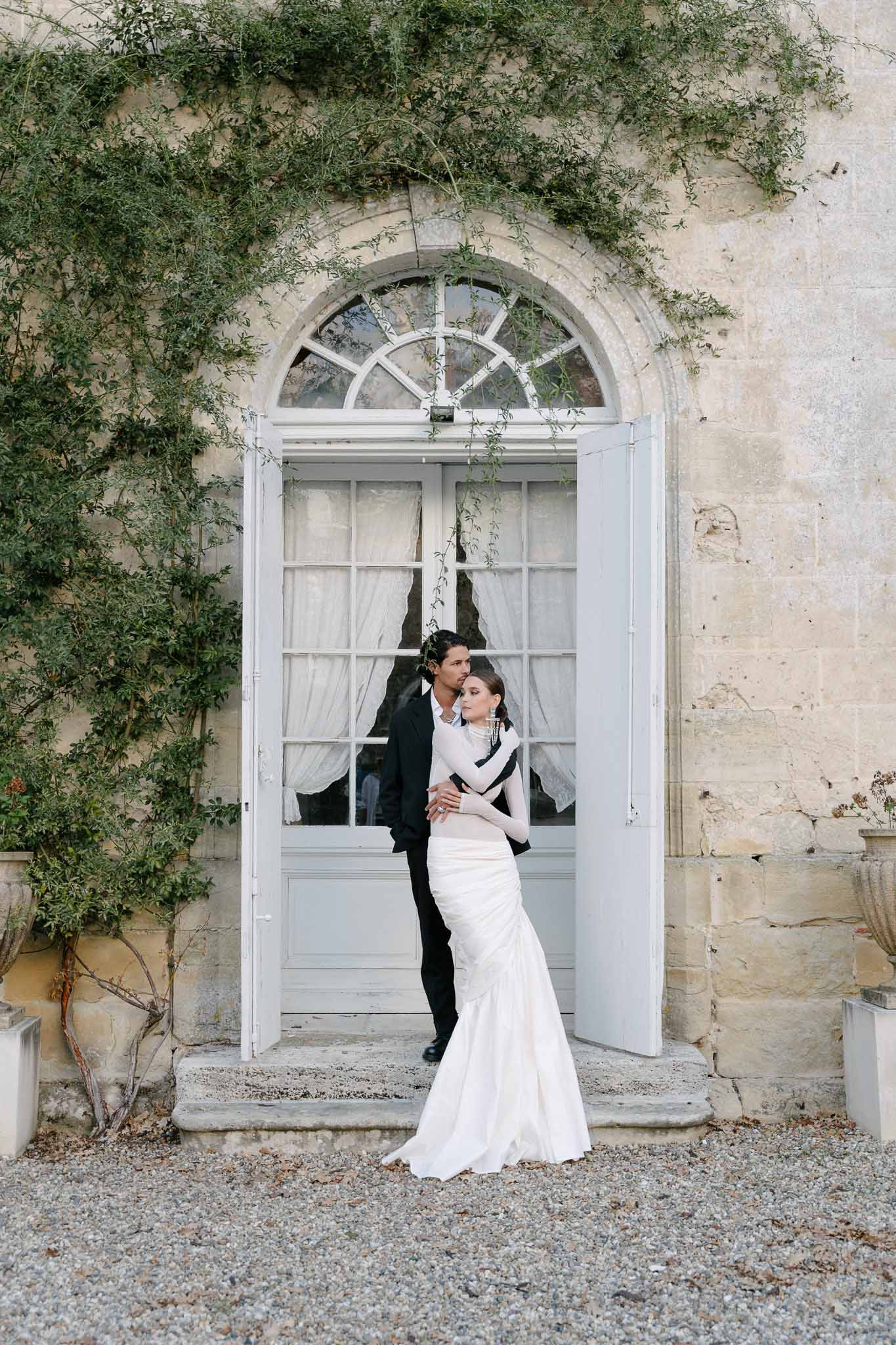 Groom embracing bride in high-neck fitted gown before pale grey arched doors on gravel courtyard