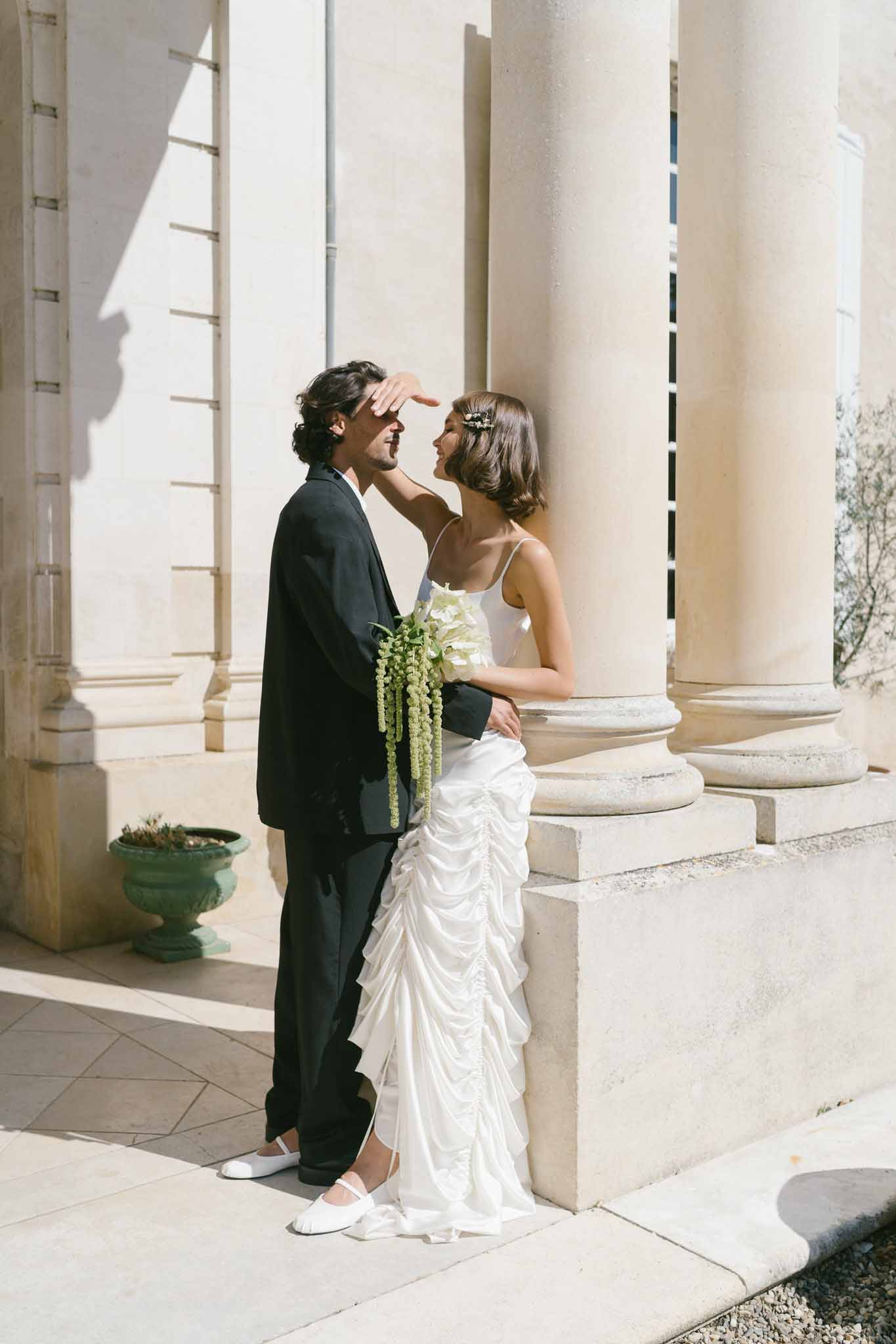 Bride playfully covering groom's eyes at stone colonnade wearing ruched satin gown with trailing green bouquet