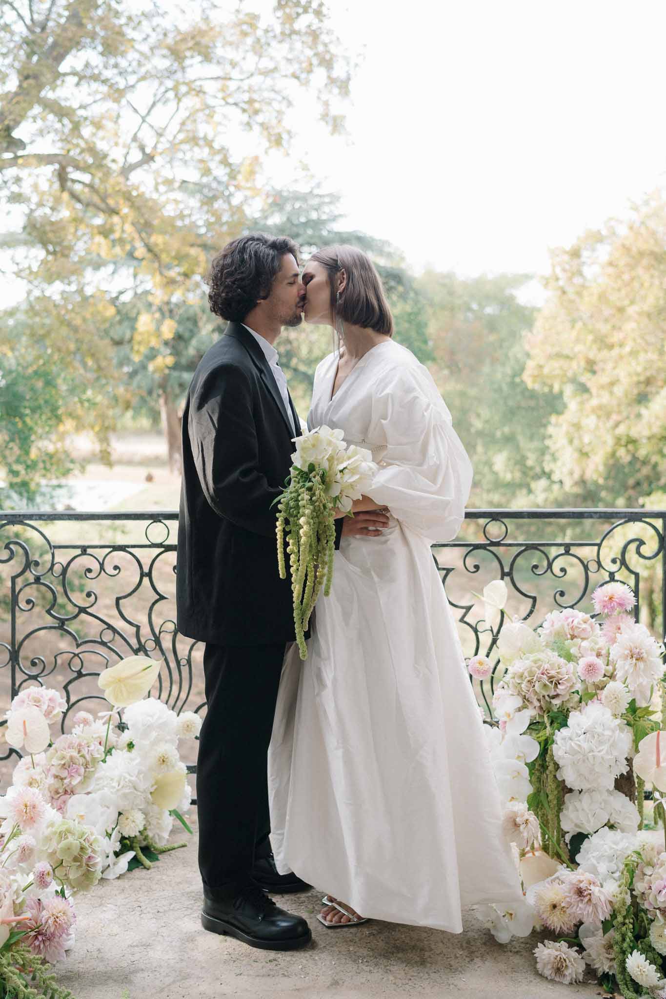 Bride and groom kissing on a terrace with wrought-iron railings flanked by white and blush floral arrangements
