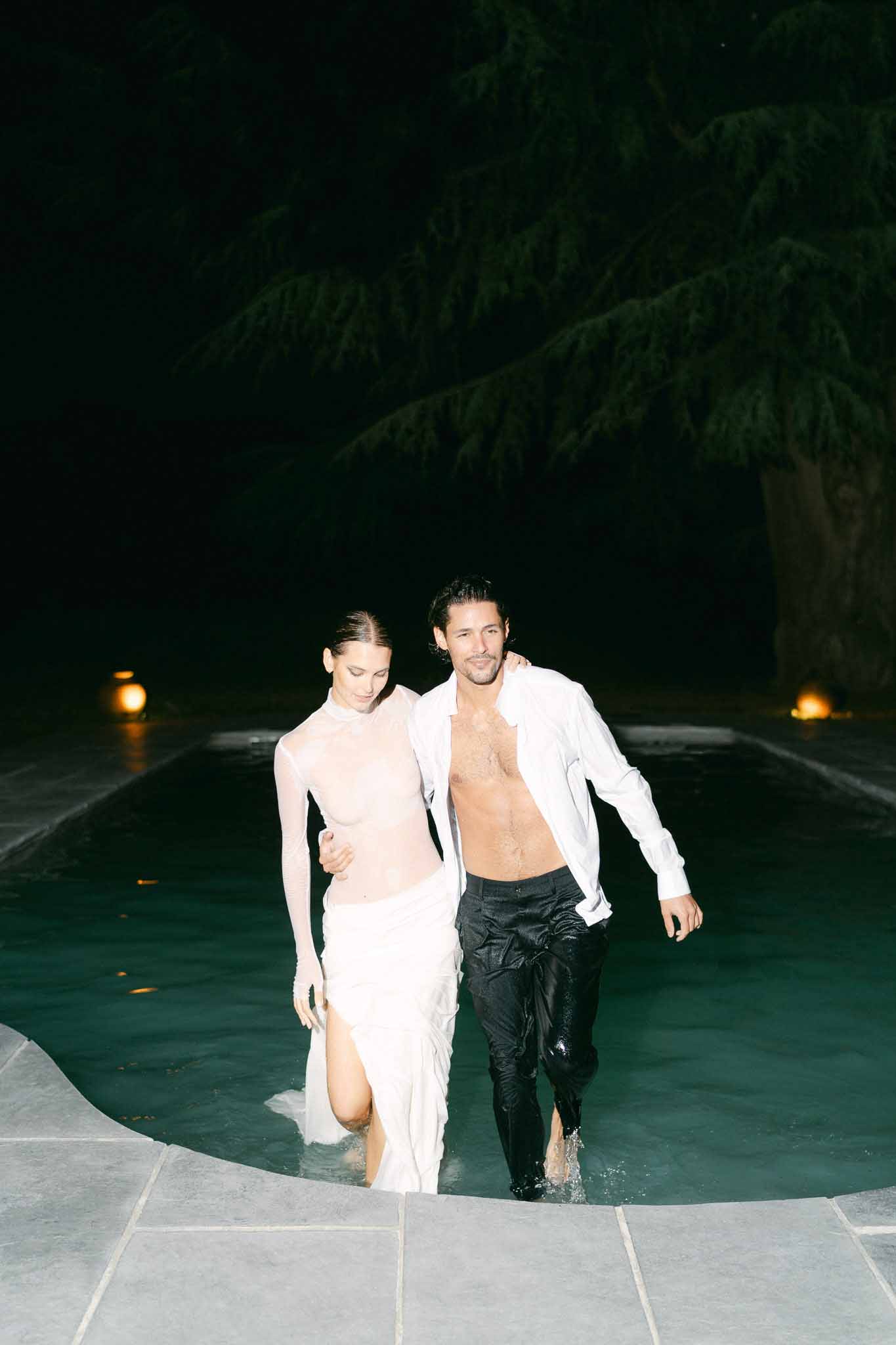 Couple wading out of illuminated pool at night in wet wedding attire