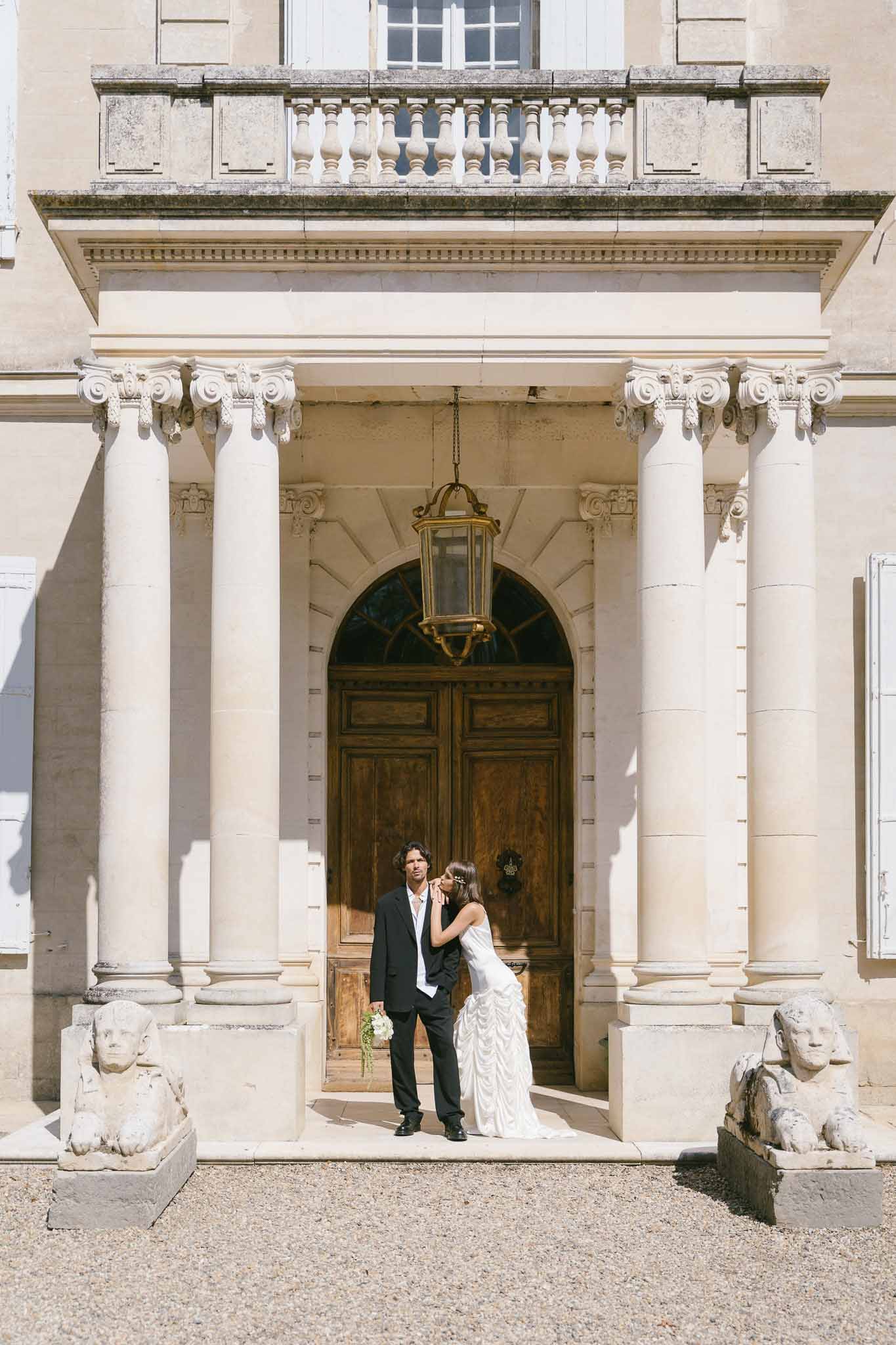 Bride kissing groom on cheek at columned chateau entrance with stone sphinx sculptures flanking the steps