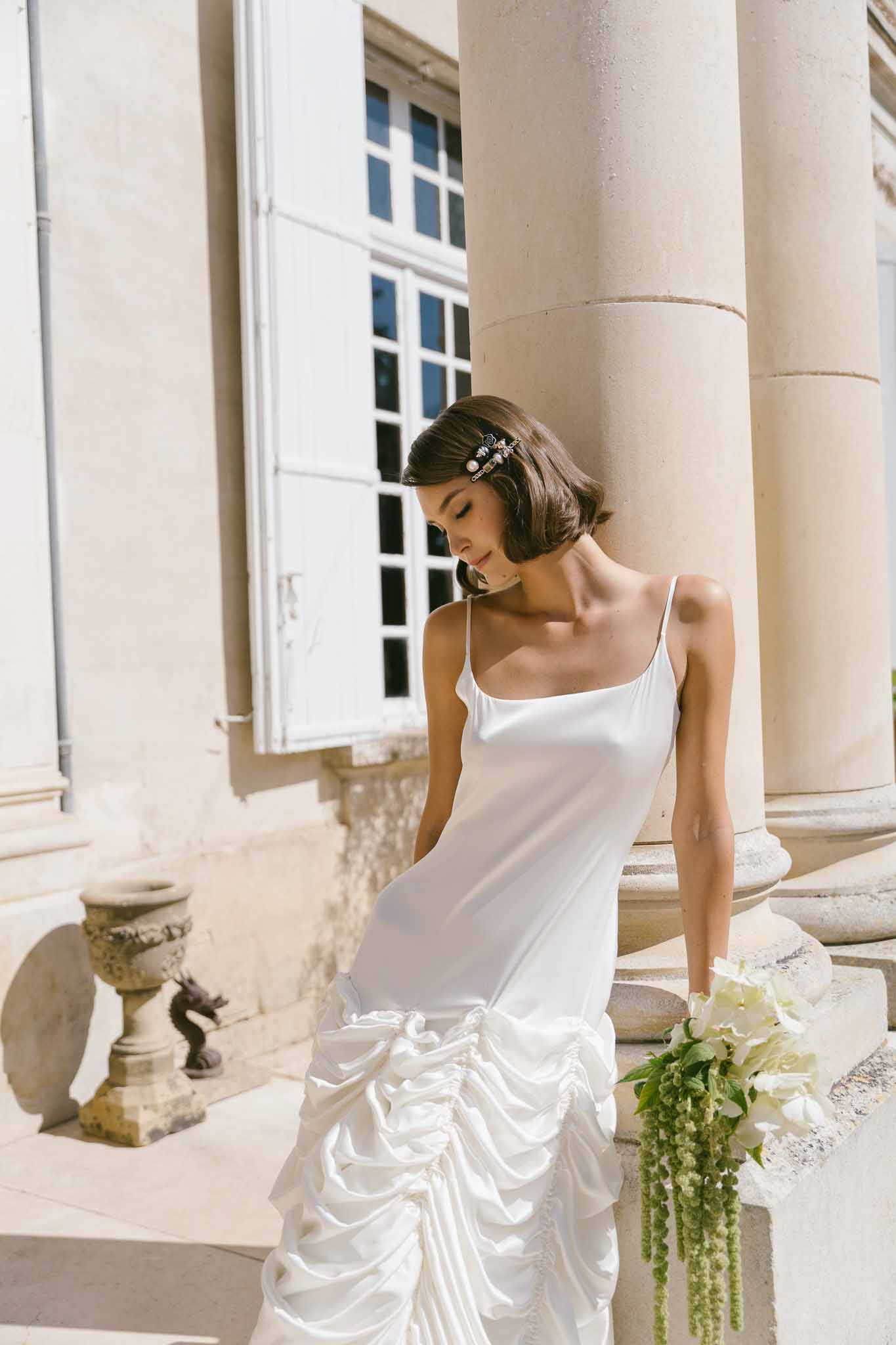 Bride with short dark bob in white satin ruched slip dress holding white hydrangea bouquet at chateau
