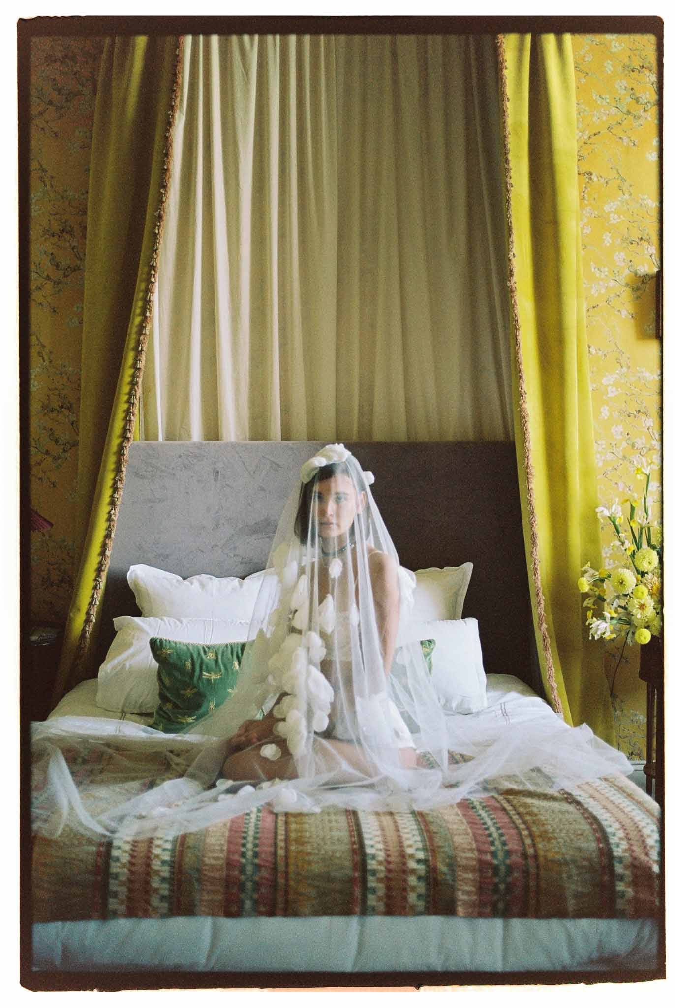 Bride in floral applique veil and flower crown sitting on four-poster bed in ornate yellow-curtained room