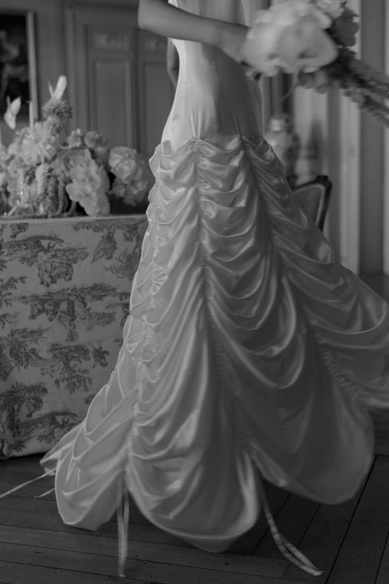 Black-and-white detail portrait of bride holding peony bouquet in draped satin gown inside a chateau room