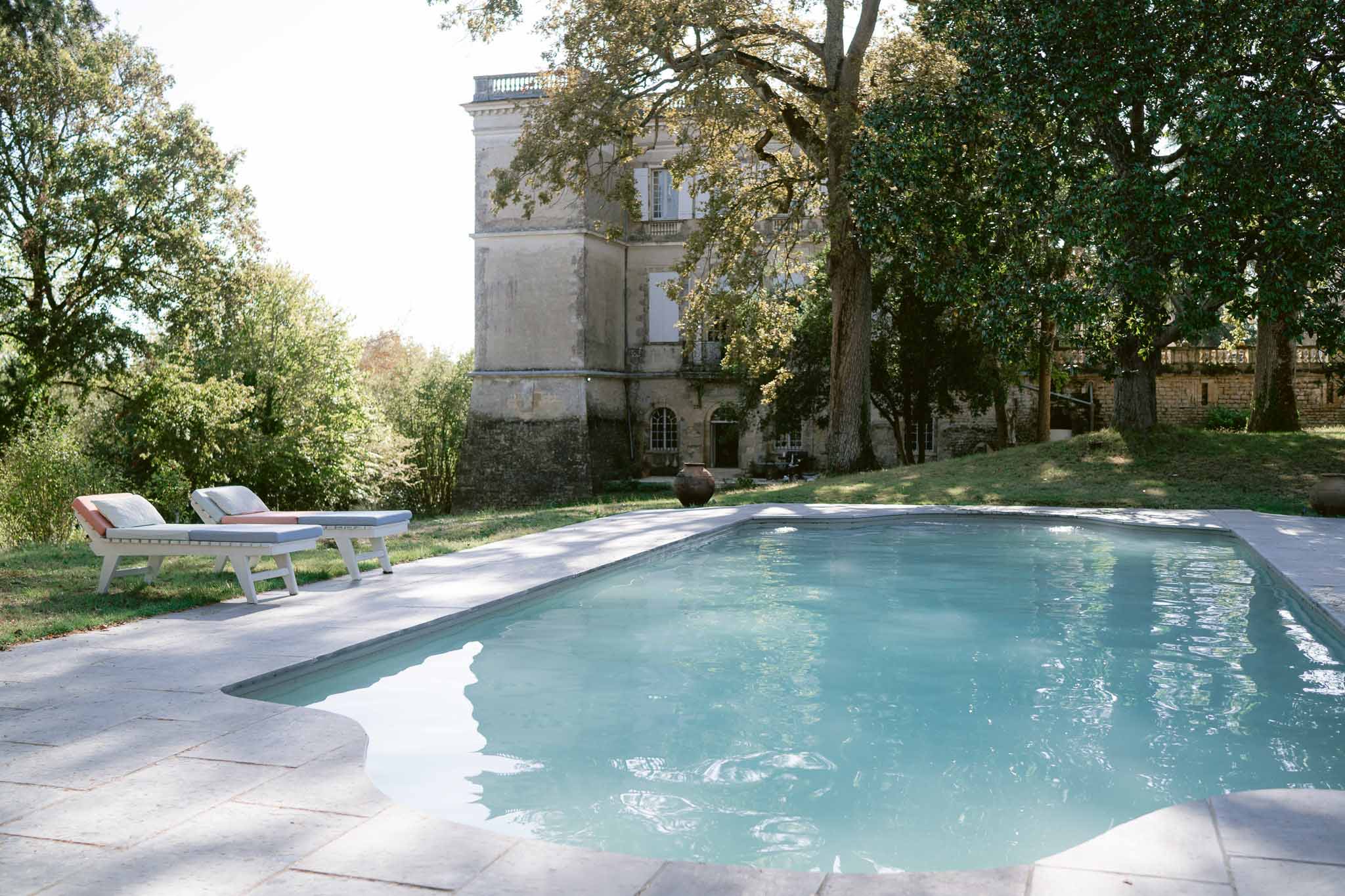 Limestone chateau with white shutters and balustraded roofline beside rectangular swimming pool