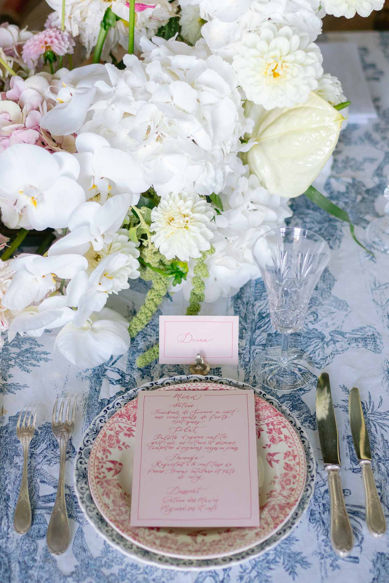 Place setting with blue toile charger, red transferware plate, calligraphy menu, and white peony centerpiece