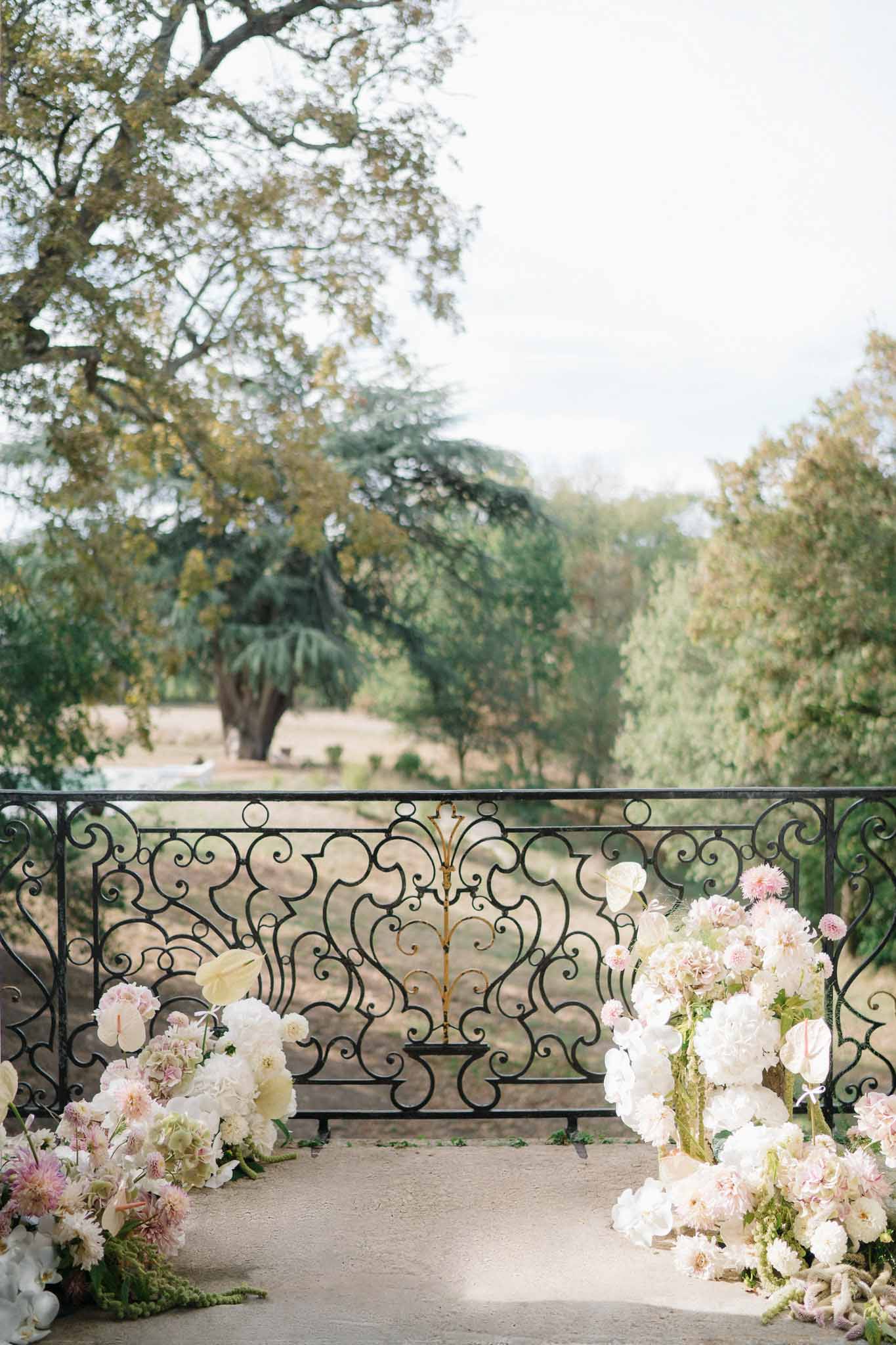White peonies, blush dahlias, and trailing green amaranthus arranged on a wrought-iron balustrade overlooking chateau grounds