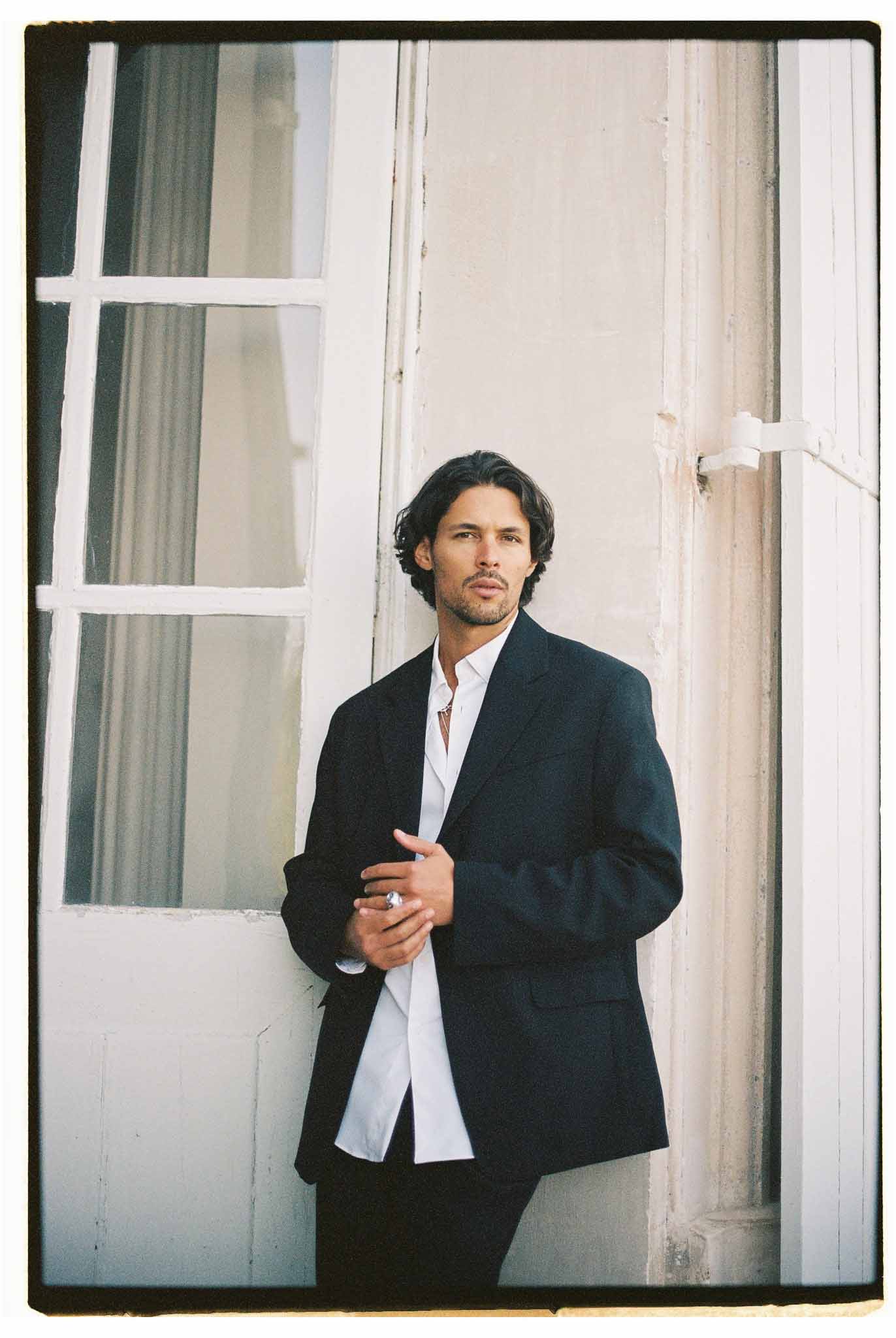 Groom in navy blazer and open-collar white shirt leaning against wall between French windows in film portrait