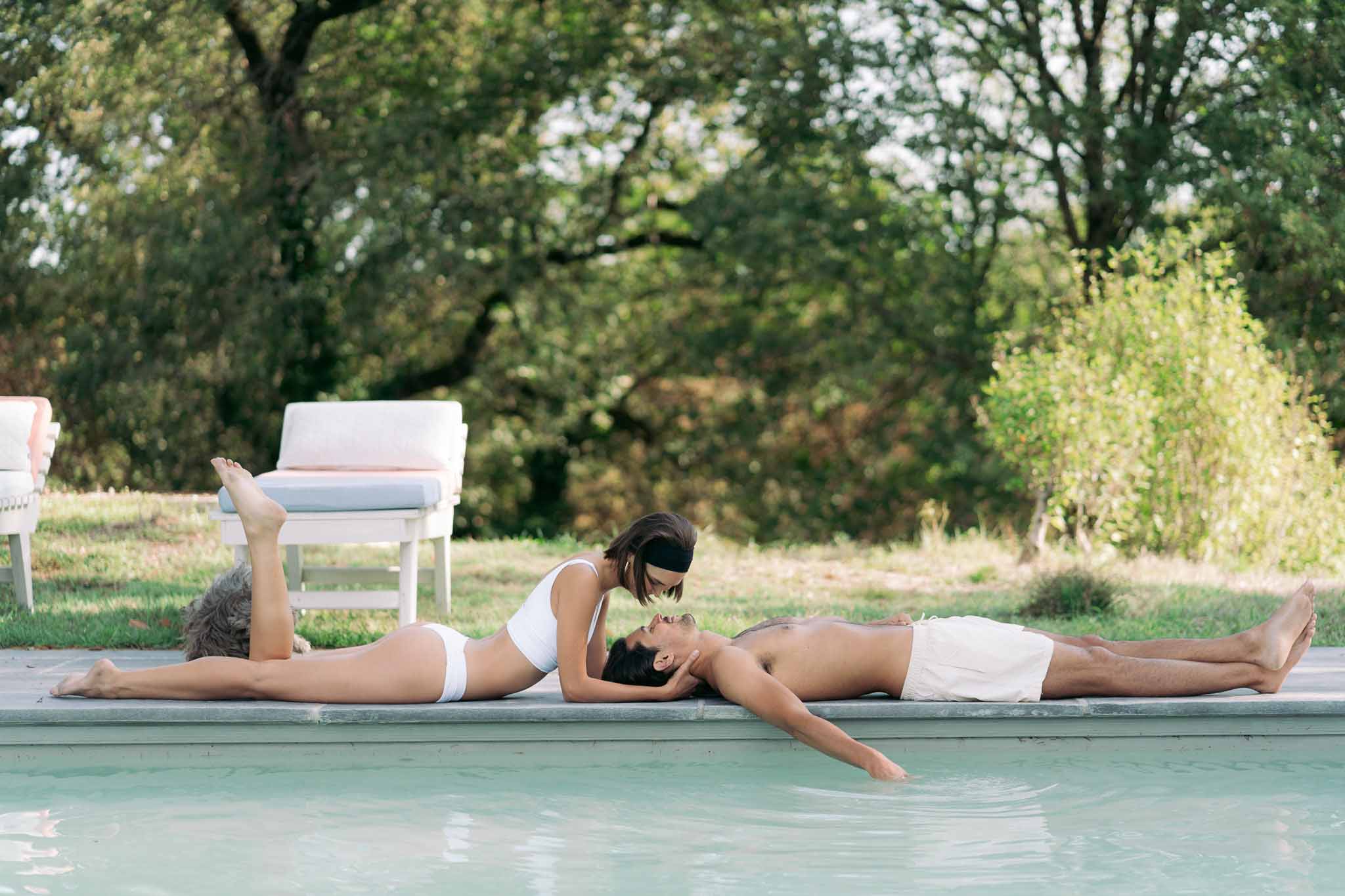 Couple lying by pool edge with white lounge chairs and pale cushions in relaxed poolside setting