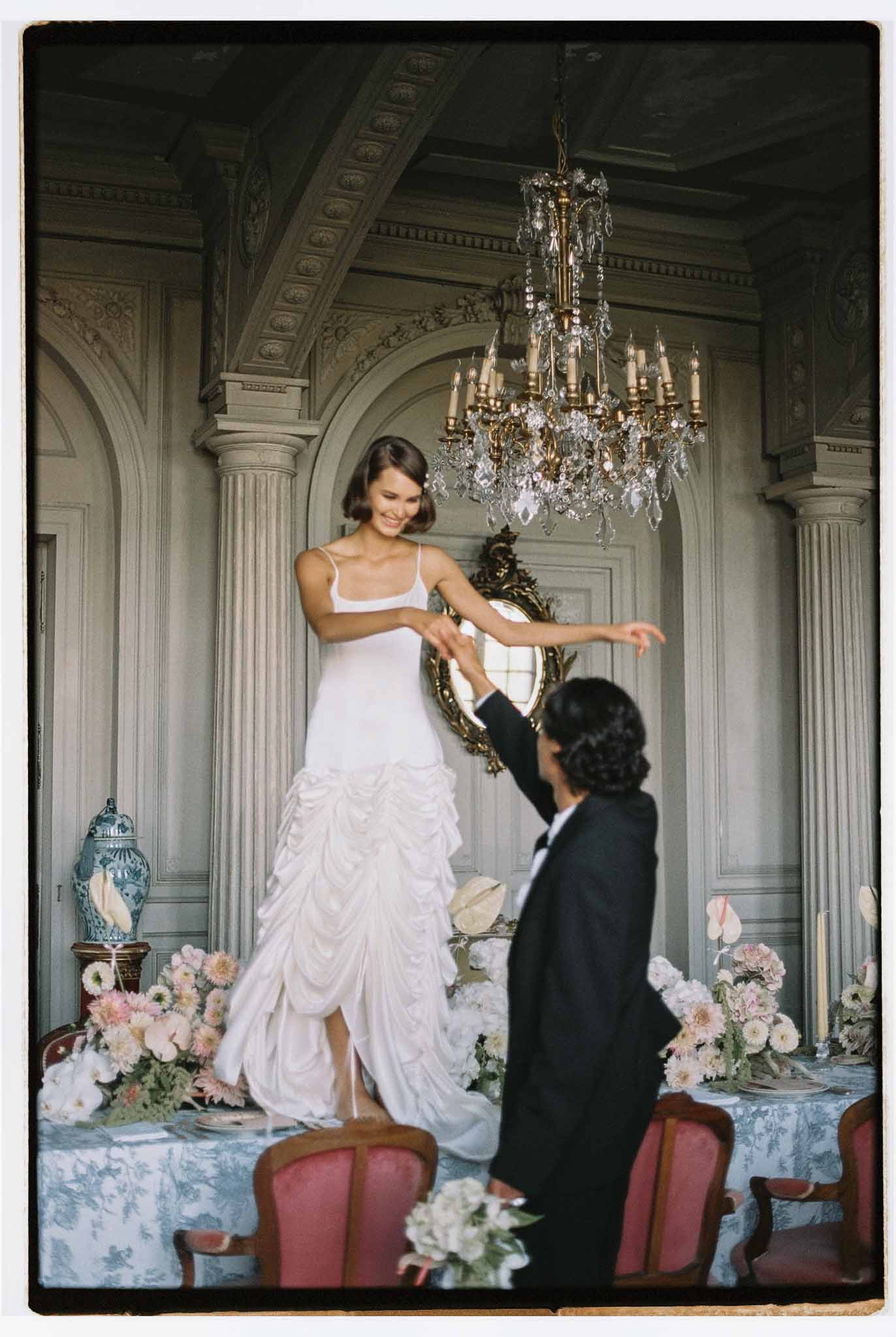 Bride standing on dining table holding groom's hand in an ornate chateau salon with chandelier and toile tablecloth