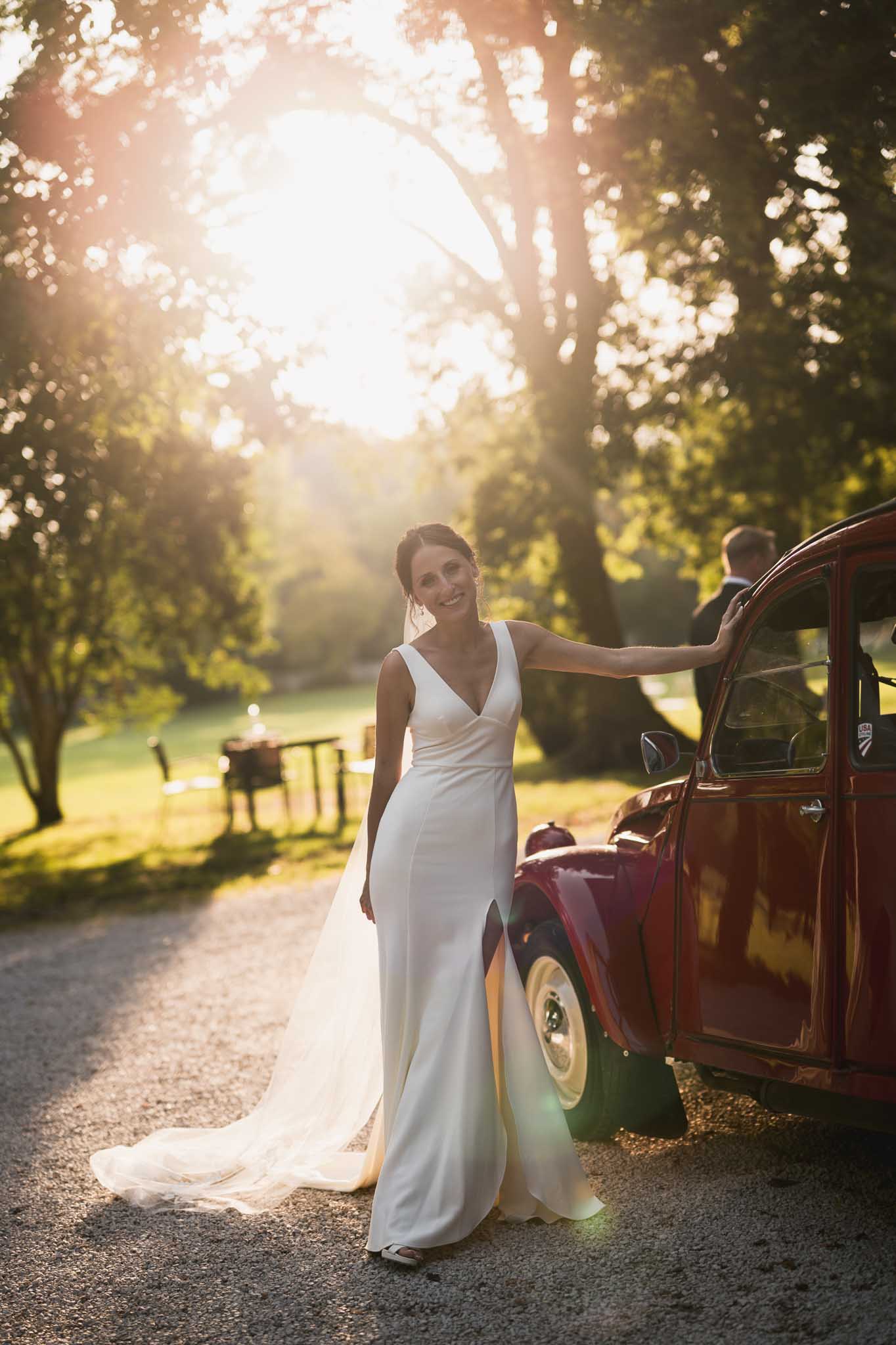 Bride in ivory gown posing beside vintage burgundy car on tree-lined country road at golden hour