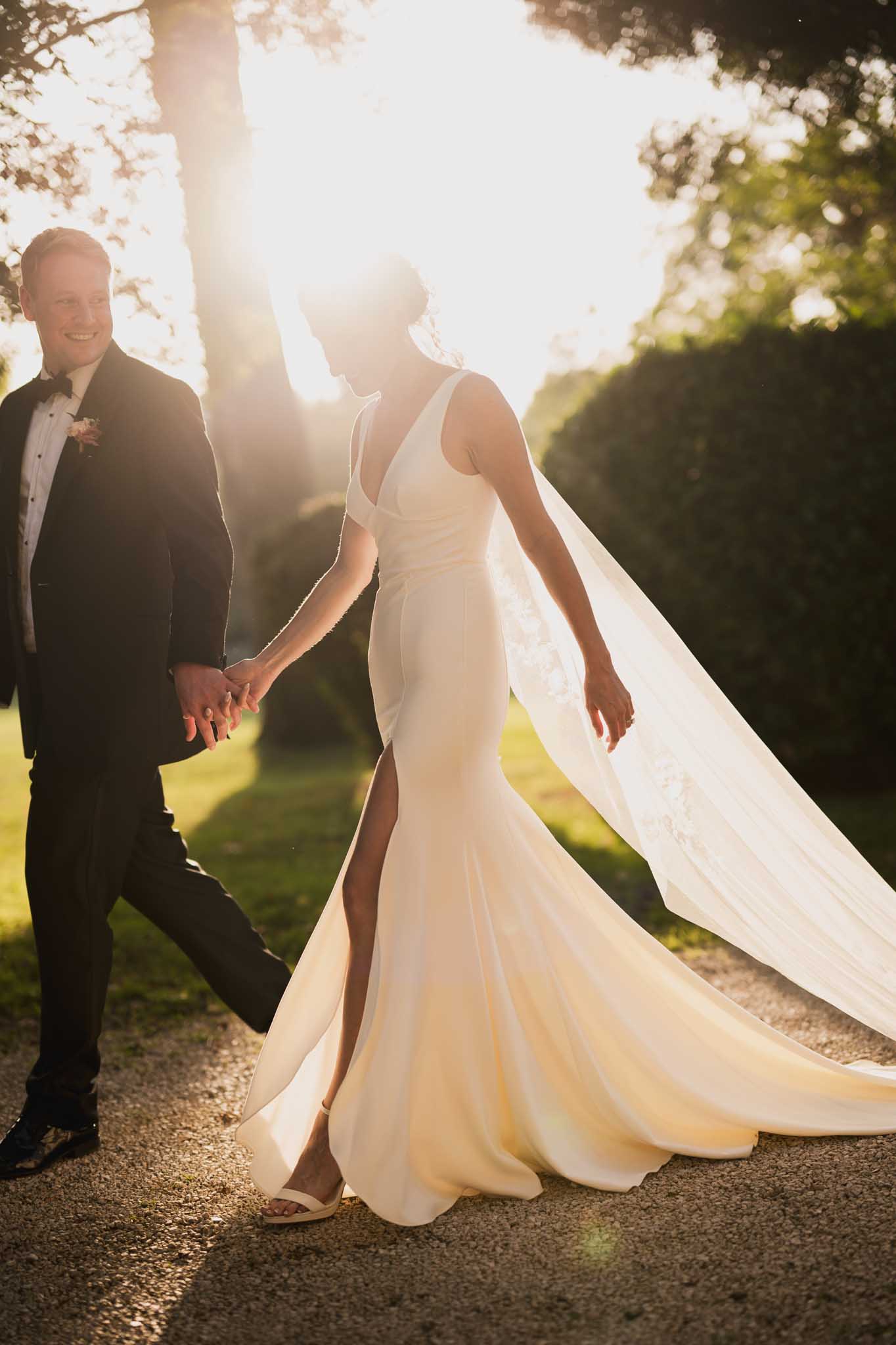Bride and groom walking hand-in-hand along tree-lined garden path during golden hour