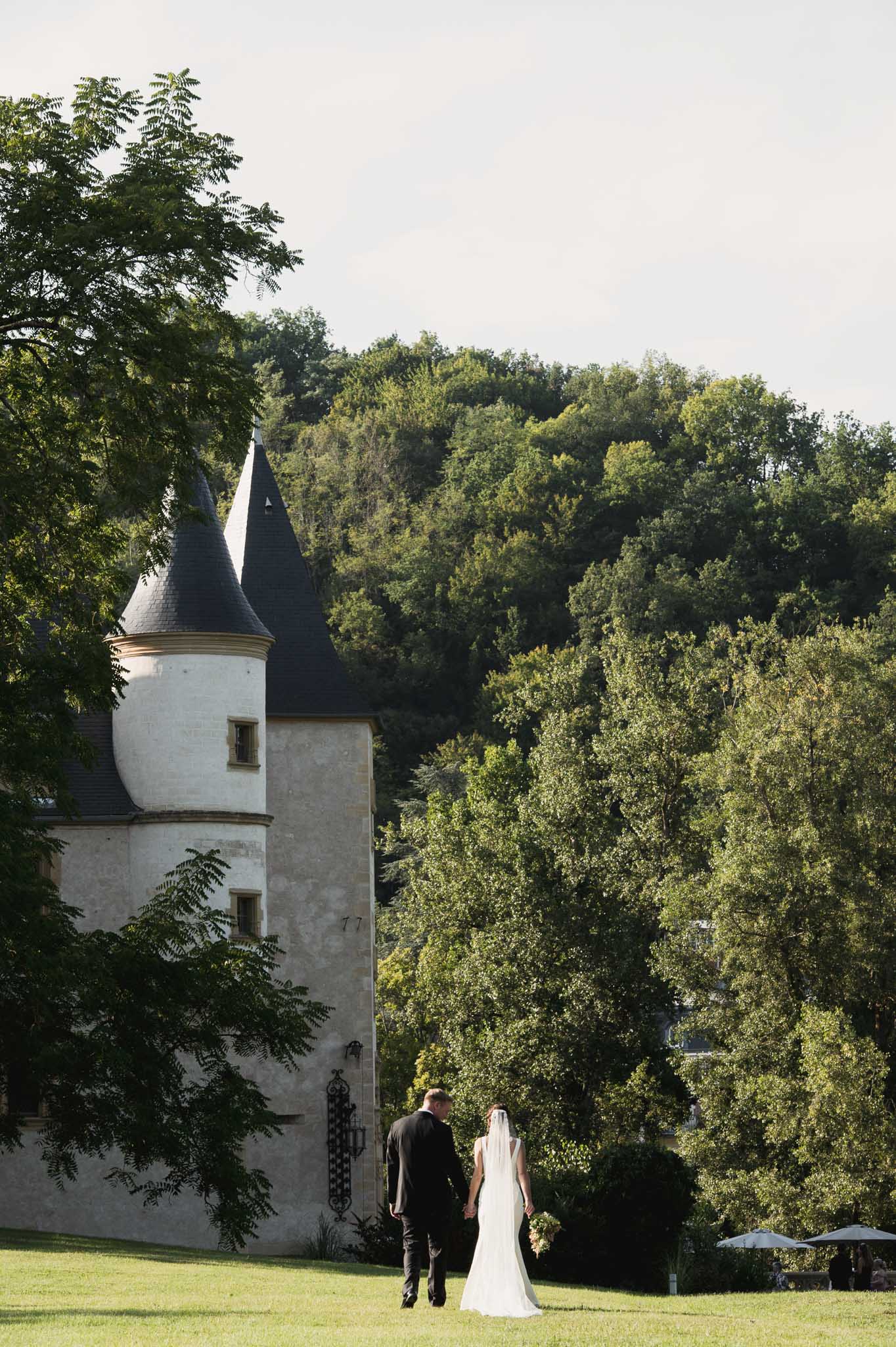 Bride and groom walking hand in hand across castle grounds with white stone tower and turrets in background