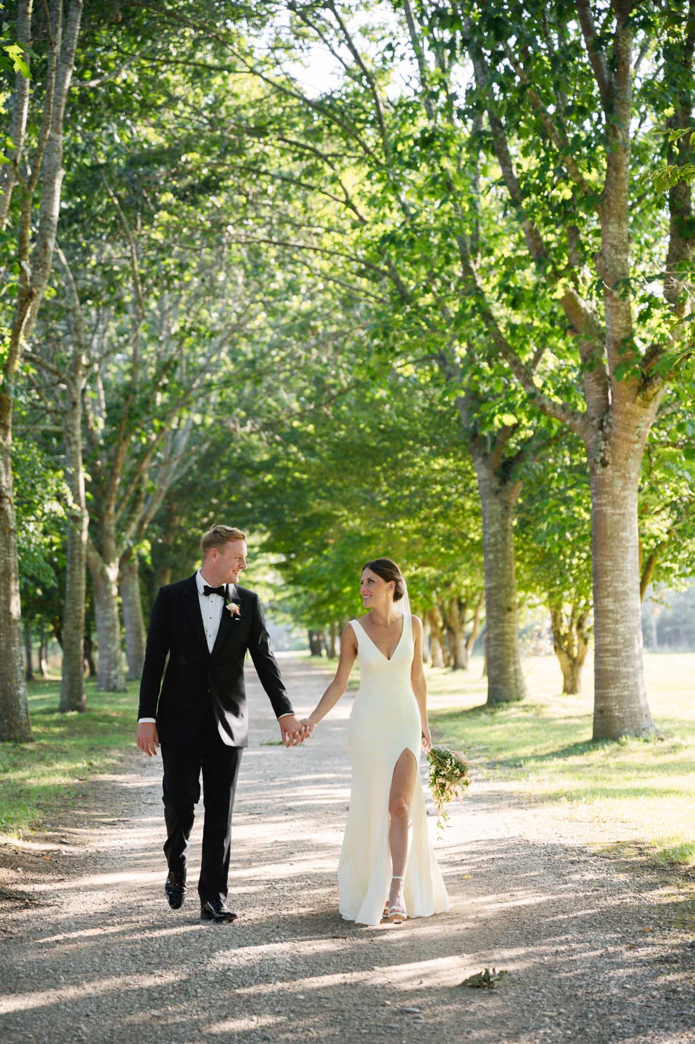 Bride and groom walking hand-in-hand down tree-lined avenue in wedding portrait