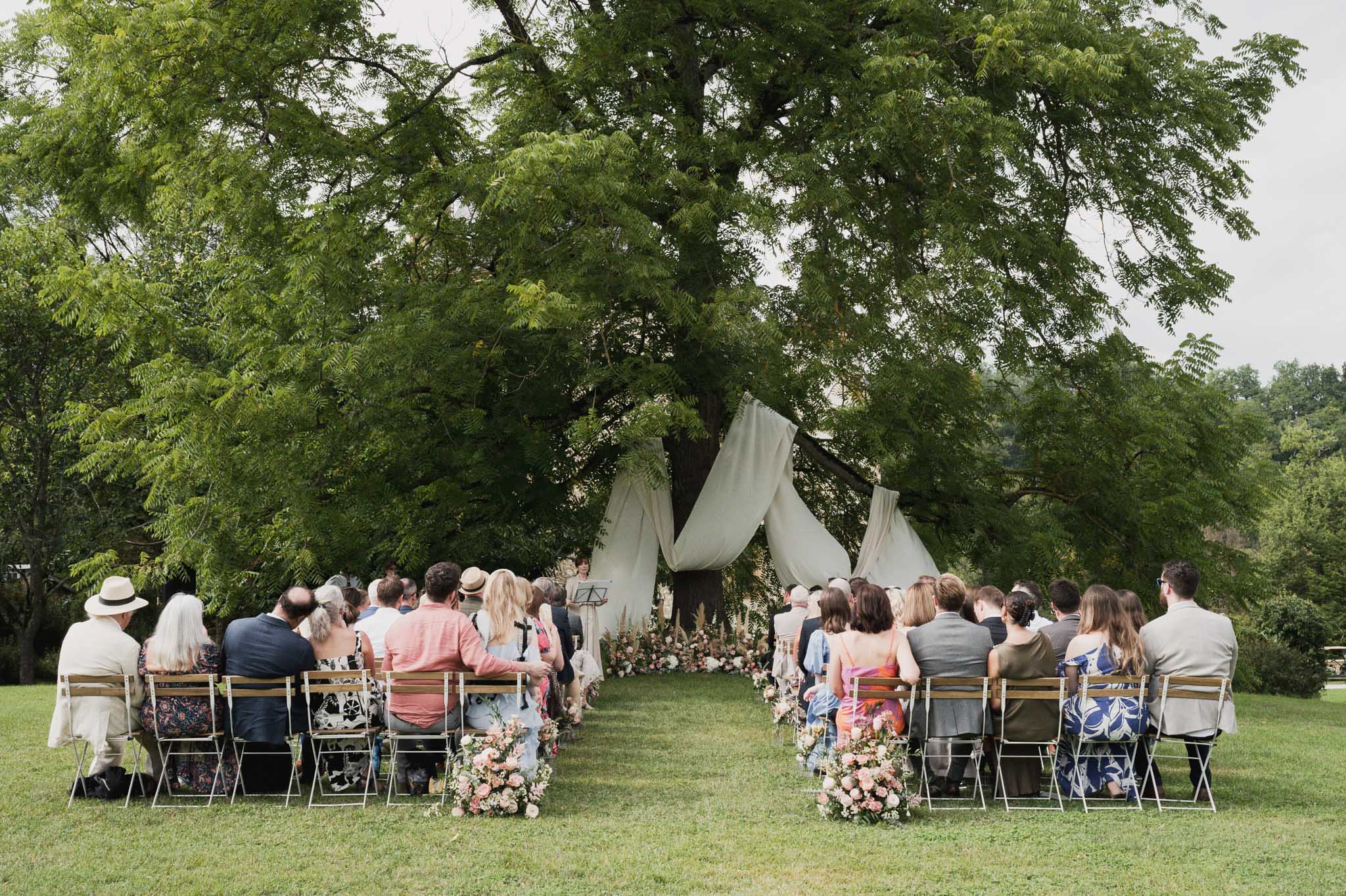 Outdoor wedding ceremony in garden setting with guests seated facing draped altar between trees