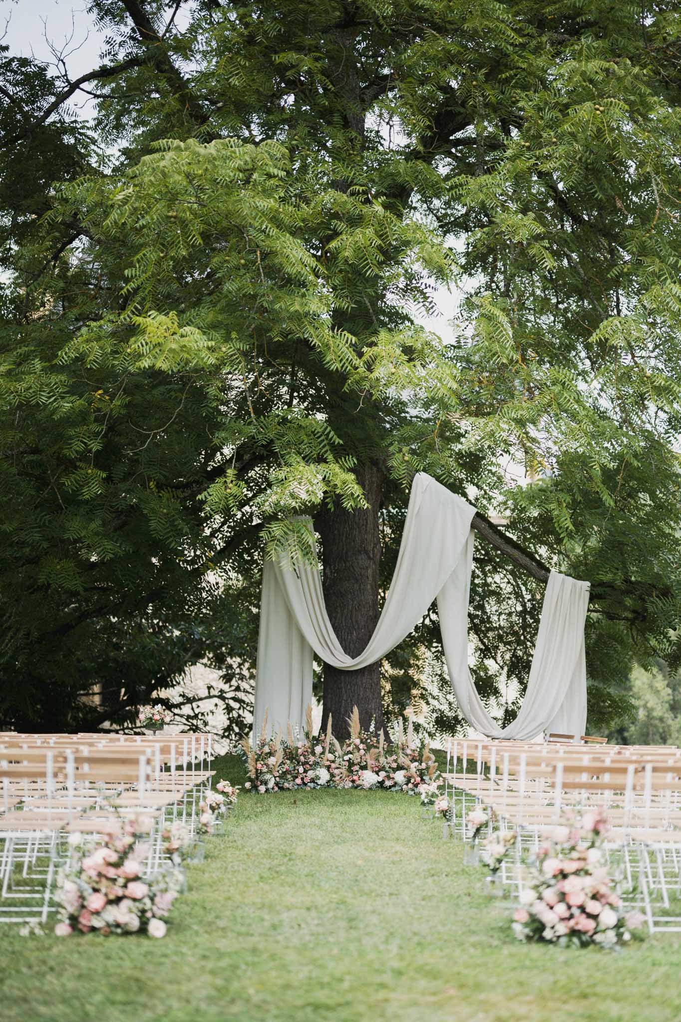 Outdoor wedding ceremony setup with ivory draping and floral aisle under mature tree in garden setting