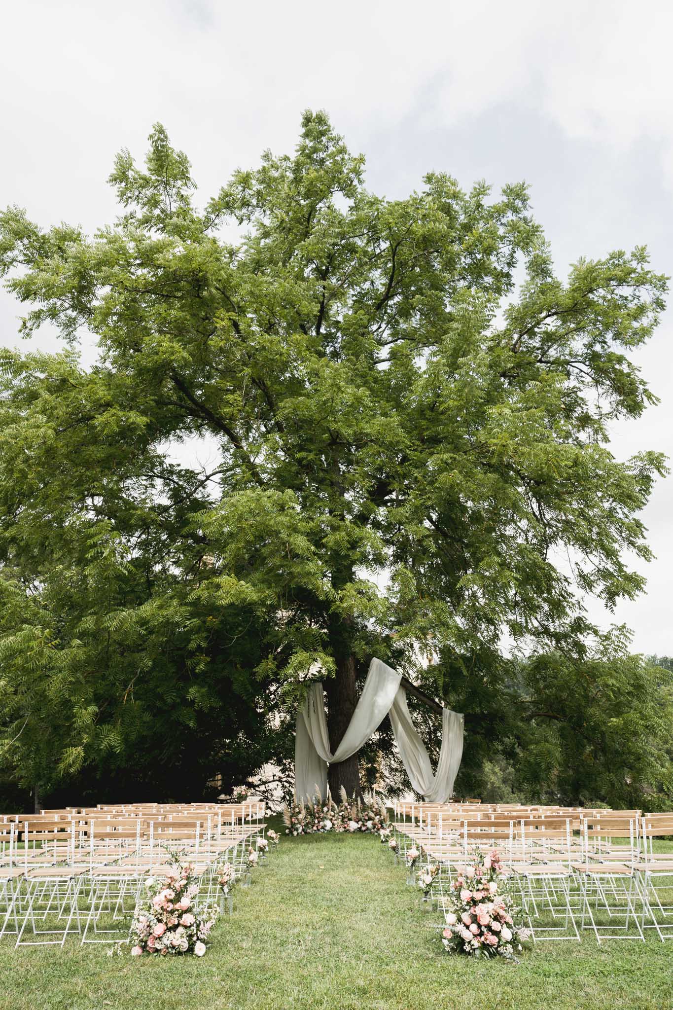 Outdoor wedding ceremony setup under tree with ivory draping and white chairs at garden venue