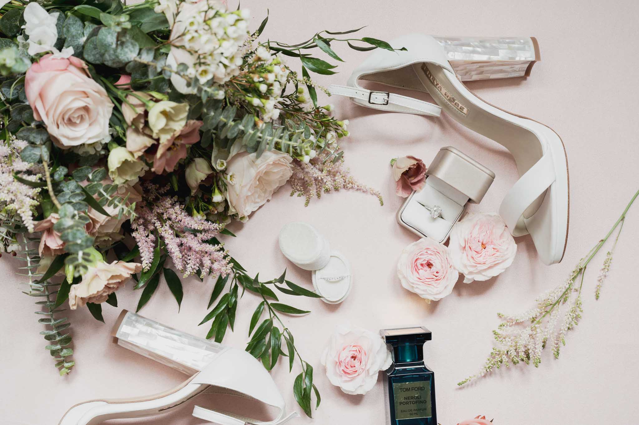 Bridal flat lay with bouquet, shoes, rings, and perfume arranged on neutral surface