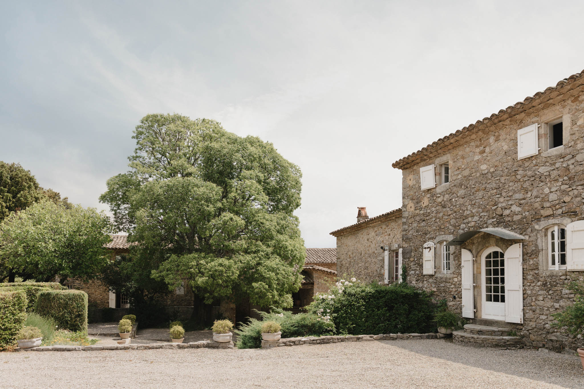 Provencal stone mas with terracotta roofs, white shutters, and gravel courtyard with clipped hedges