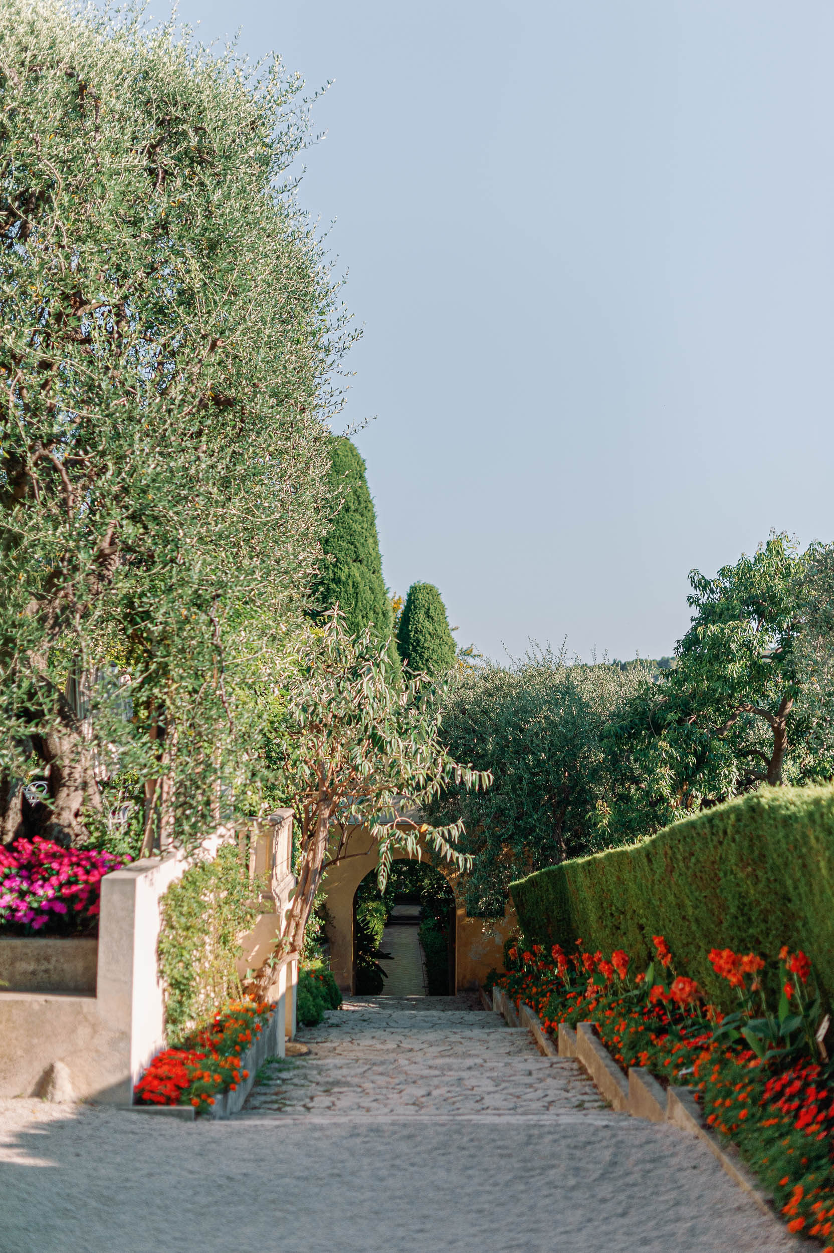 Cobblestone garden path leading to an ochre archway flanked by olive trees and flowering plants at a Provencal estate