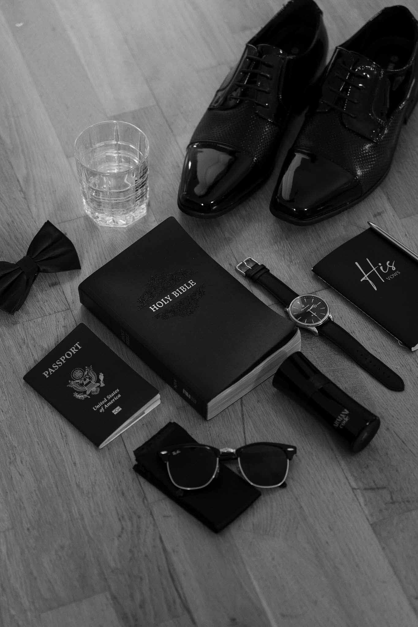 Black and white flat lay of groom accessories with Oxford shoes, bow tie, watch, and vow booklet
