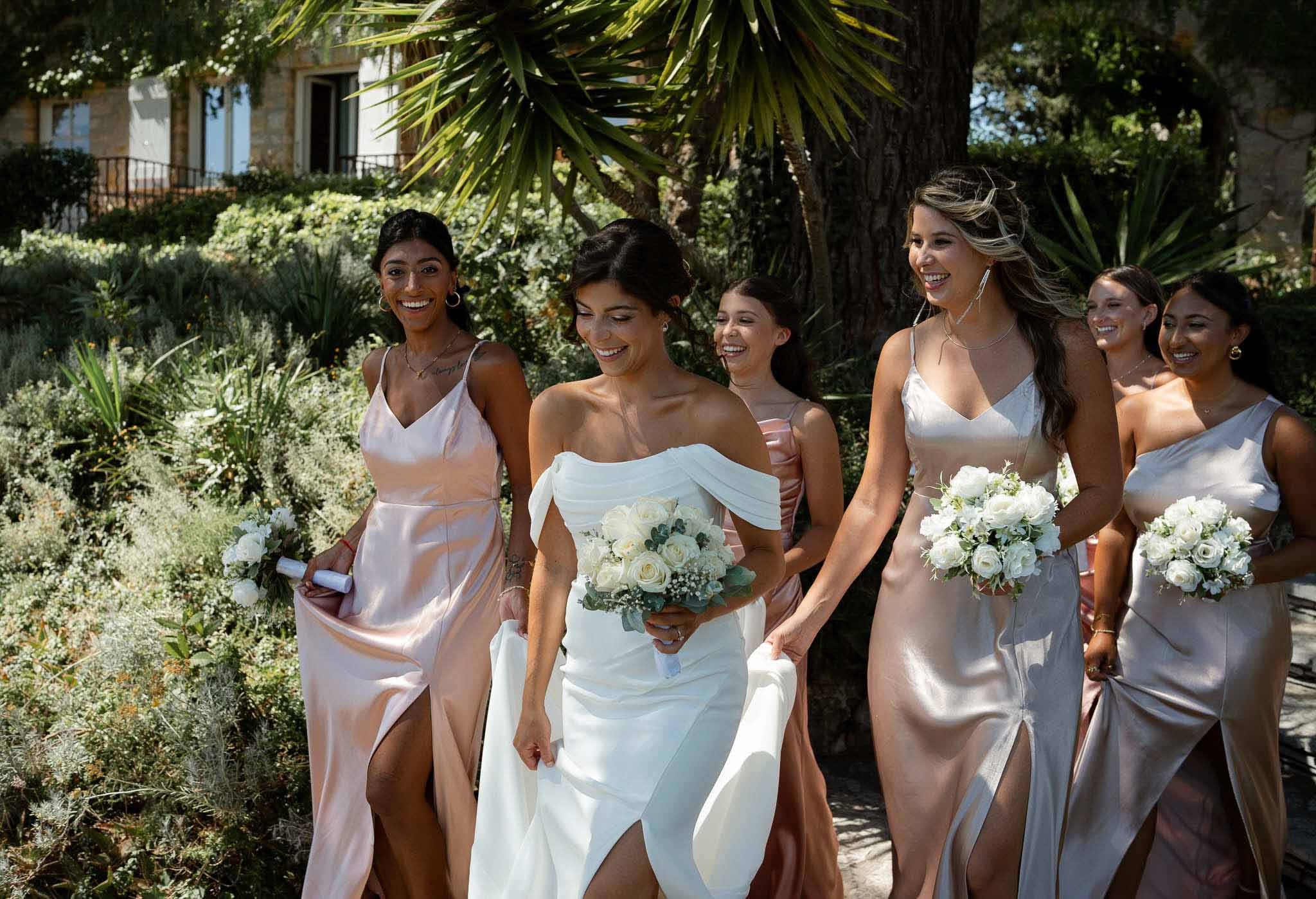 Bride and five bridesmaids in mismatched satin slip dresses walking and laughing on chateau grounds