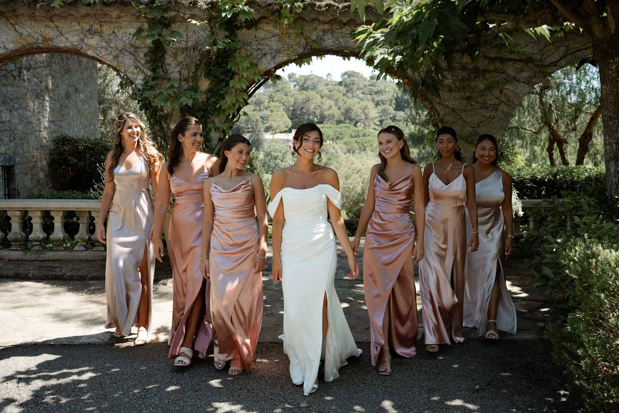 Bride and six bridesmaids in blush and rose gold satin dresses walking under ivy-covered stone arches at a French chateau
