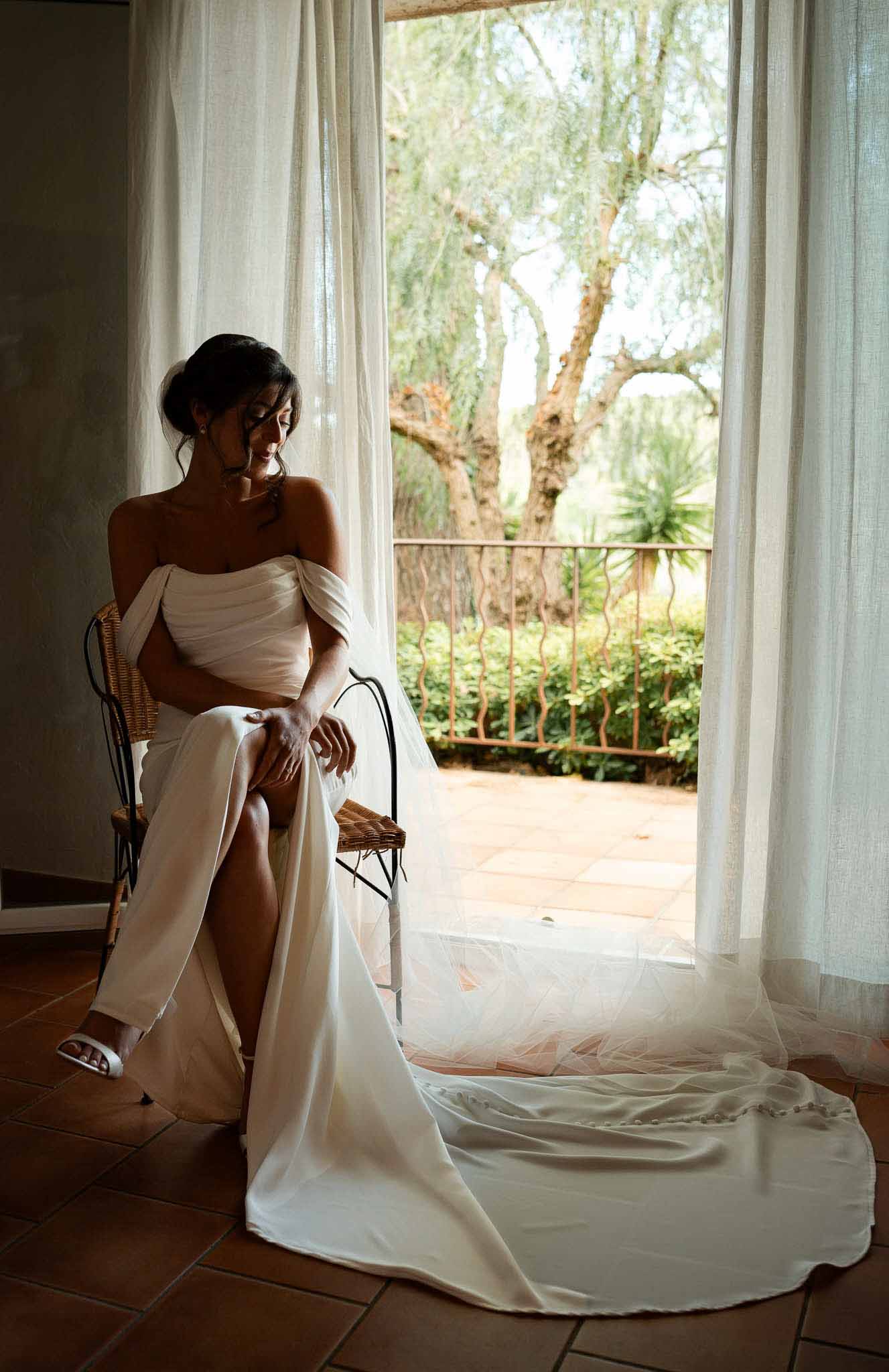 Bride in off-shoulder gown with cathedral train seated by open window with wrought-iron terrace beyond